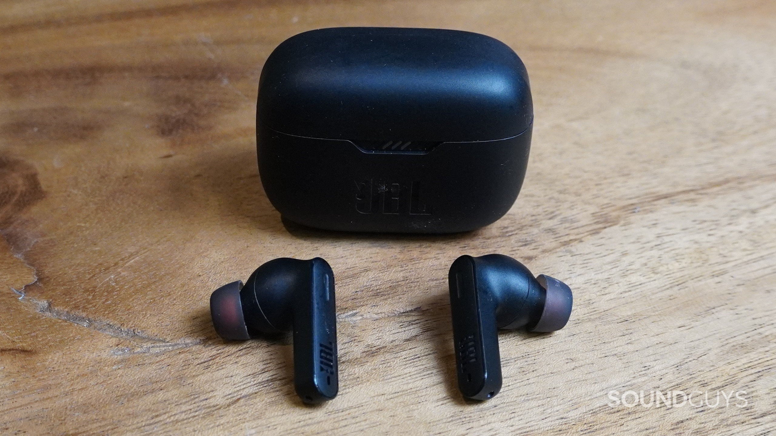 The JBL Tunes 230NC TWS earbuds shown outside and next to the charging case on a wooden surface.