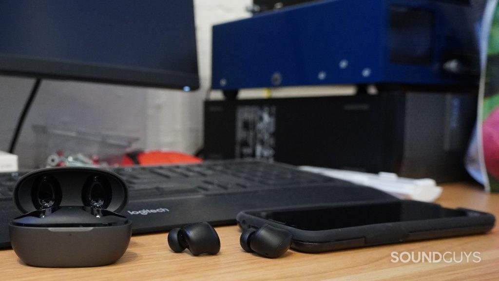 The 1MORE ColorBuds 2 lying next to its open charging case and a phone in front of a keyboard and monitor.