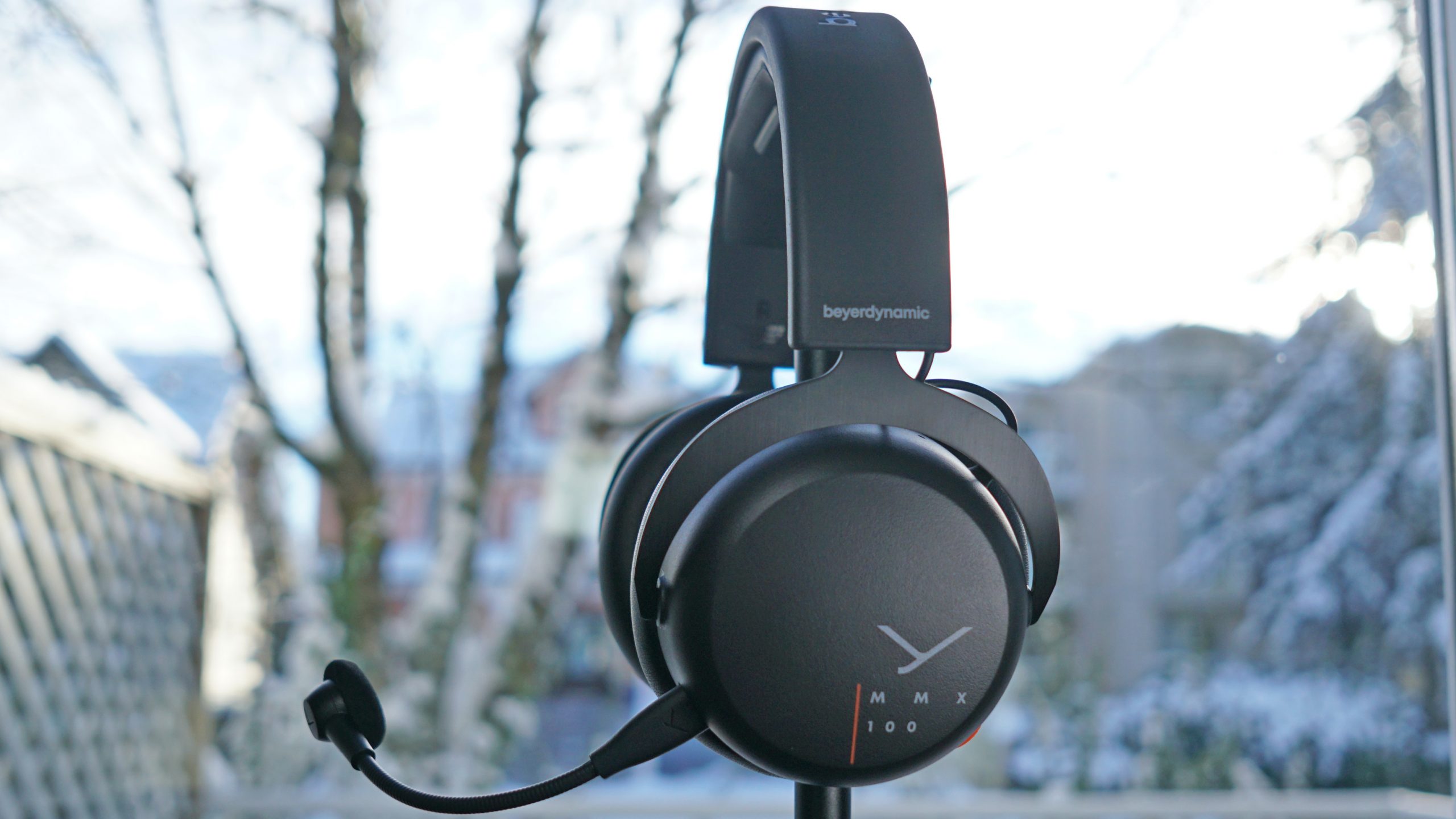 The Beyerdynamic MMX 100 gaming headset sits on a headphone stand in front of a window.