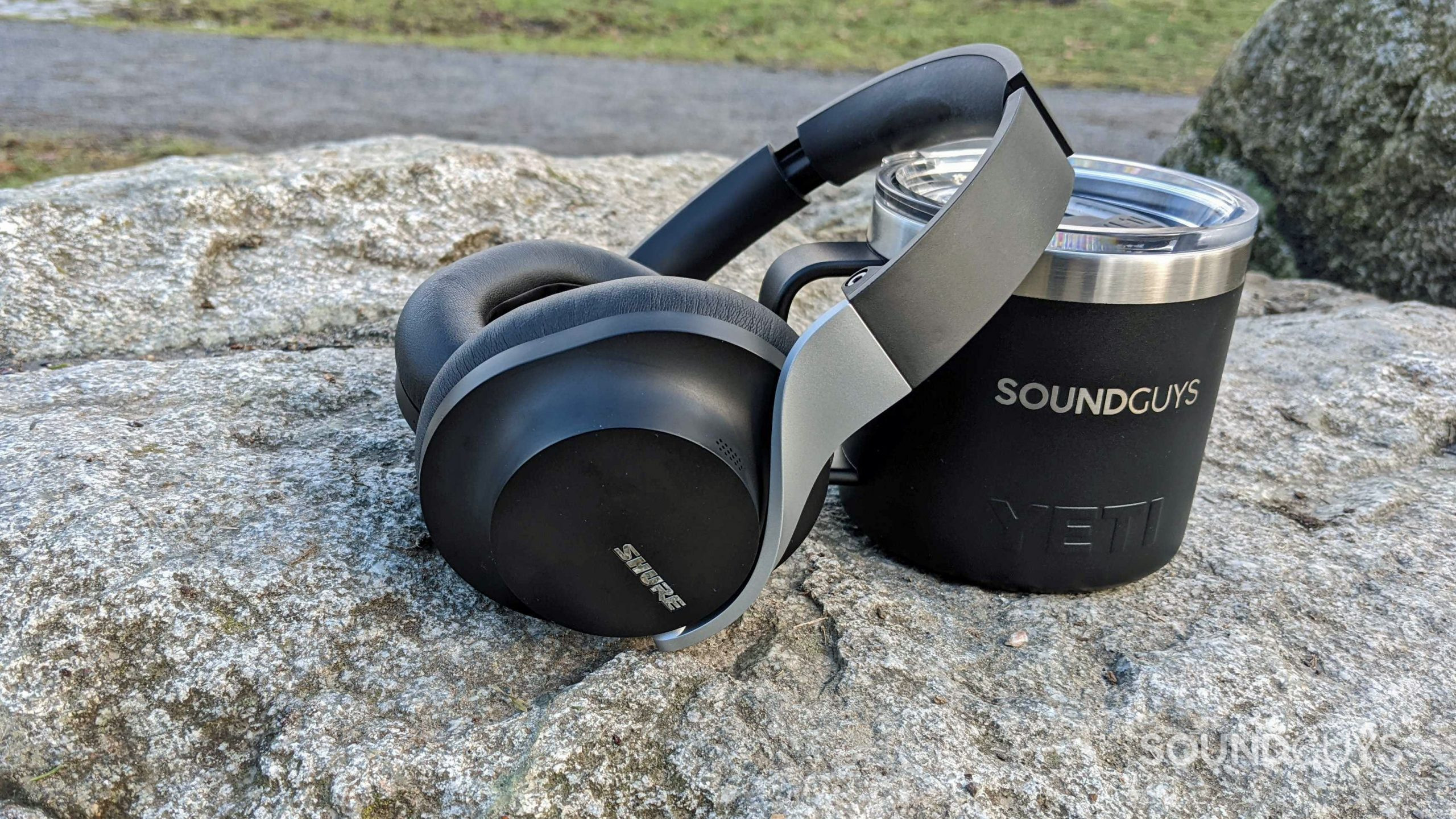The Shure AONIC 40 sits on a rock outside, leaning on a SoundGuys branded YETI travel mug.