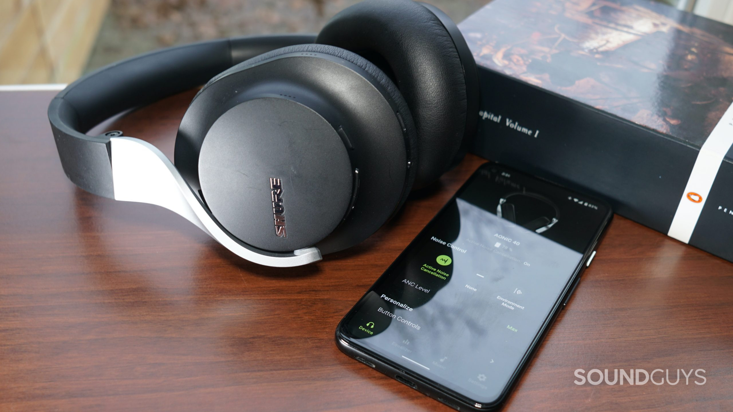 The Shure AONIC 40 bluetooth headphones lay on a wooden table next to a Google Pixel 4a running the Shure Plus Play app and a copy of volume 1 of Capital by Karl Marx.