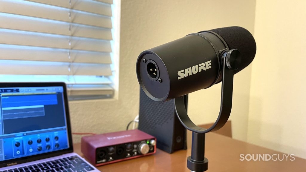 Shure MV7X showing the XLR port. A laptop, audio interface, and WiFi modem are visible in the background.