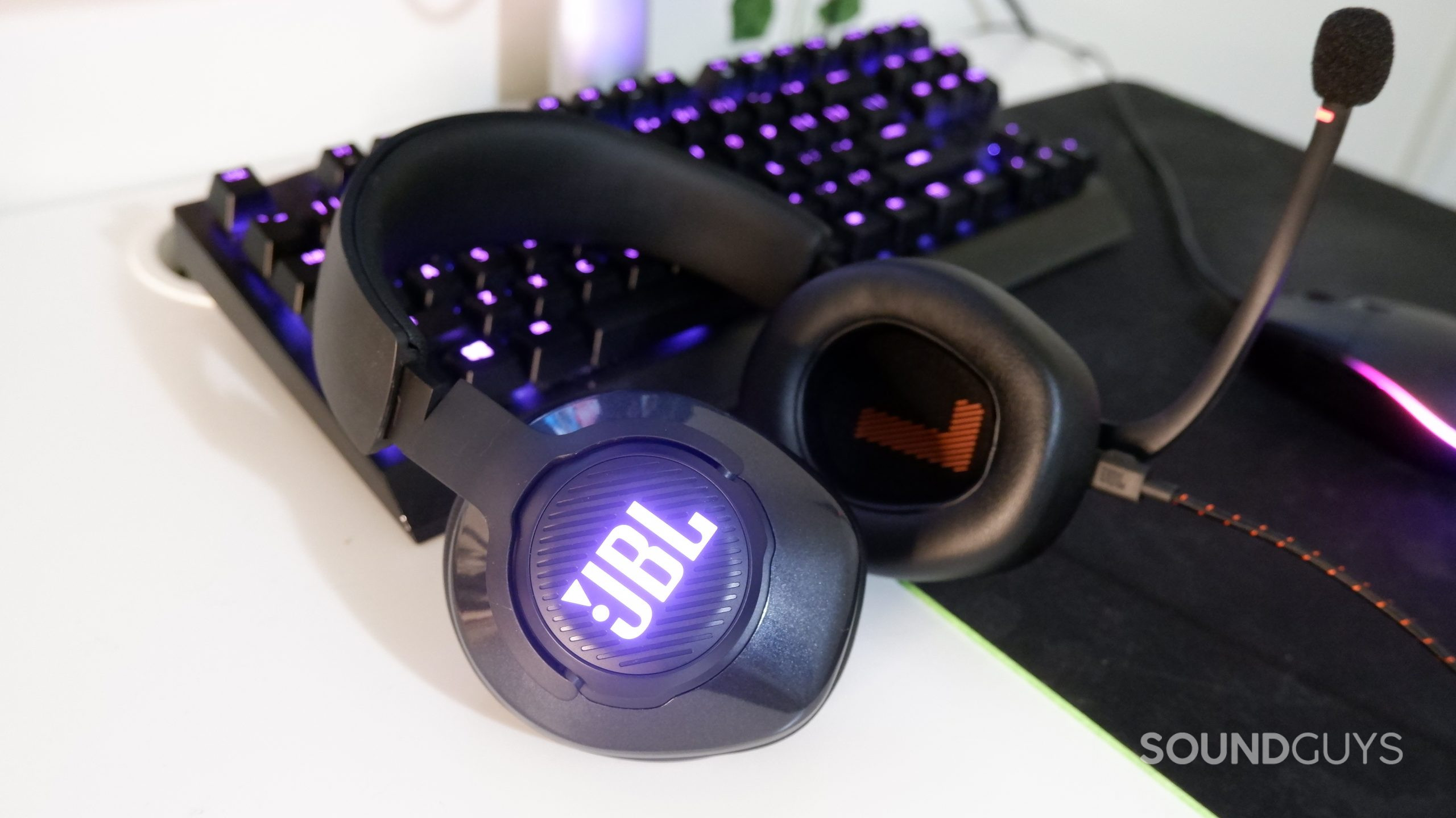 The JBL Quantum 400 laying on a white desk against an RGB keyboard, both lit up with magenta lights.