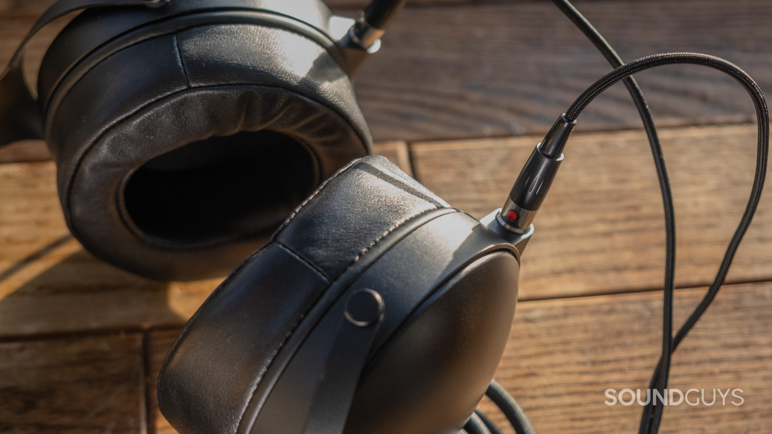 The Monolith by Monoprice M1570C sits on a wood surface with focus on the metal ear cup, leather pads, and cable.