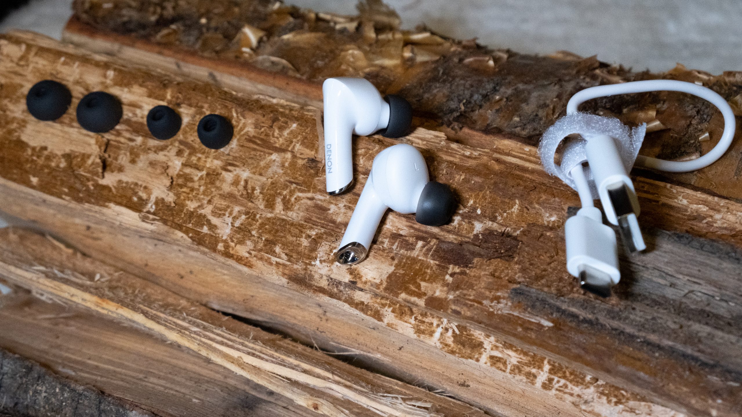 The earbuds and two extra sets of ear tips alongside included USB-C cable included with the Denon AH-C830CNW rest on a piece of rough wood.