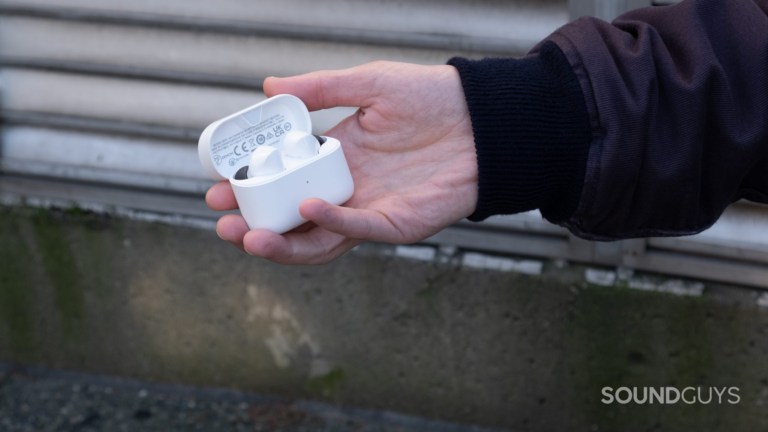 A hand holds the open case of the Denon AH-C830CNW outside by concrete and metal vents.