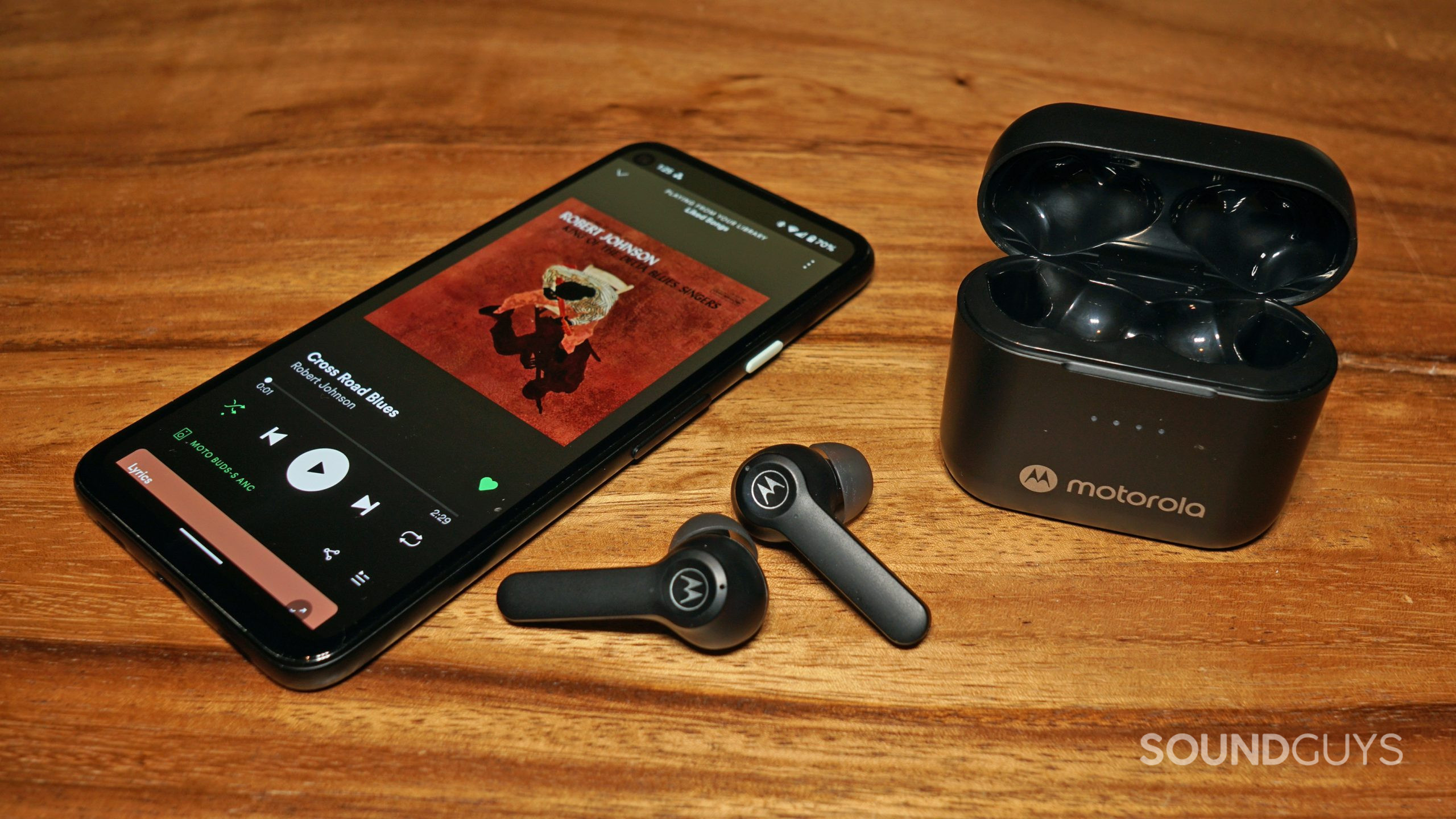 The Motorola Moto Buds S ANC lays on a wooden table with the earbuds next to the case and a Google Pixel 4a running Spotify with Cross Road Blues by Robert Johnson playing.