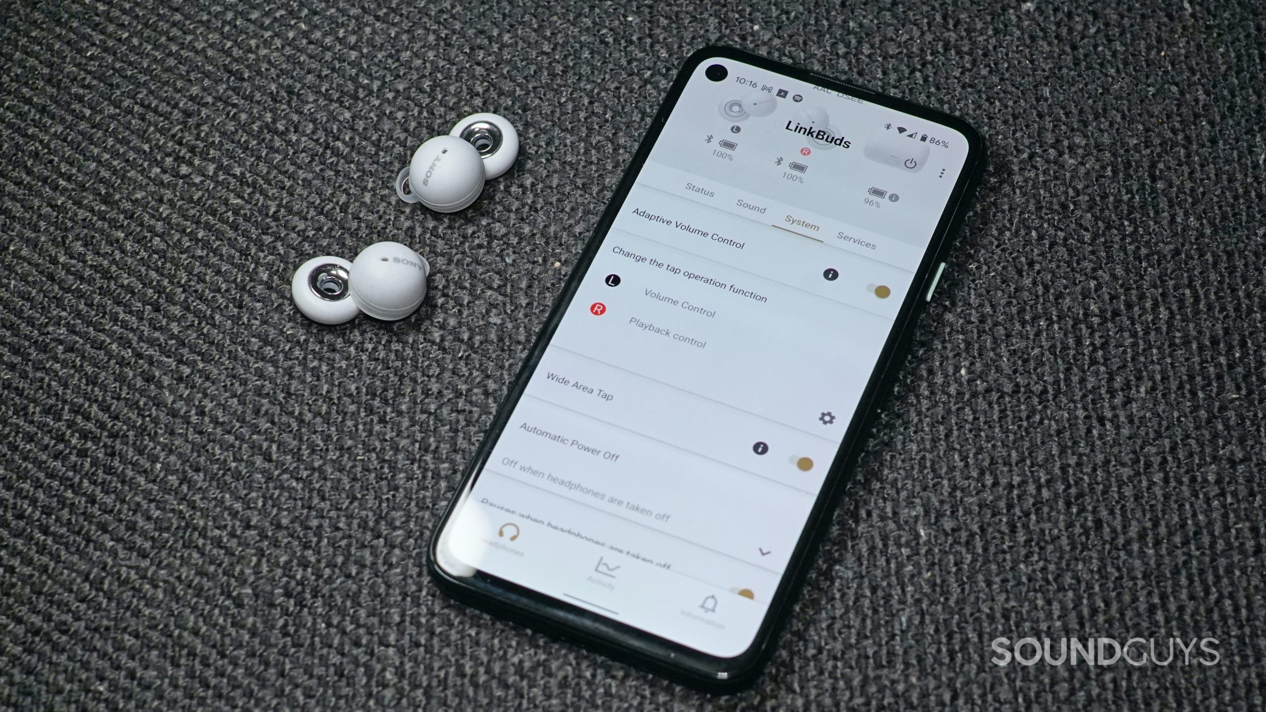 The Sony LinkBuds lie on a fabric surface next to a Google Pixel 4a displaying the Sony LinkBuds page of the Sony Headphones Connect app.