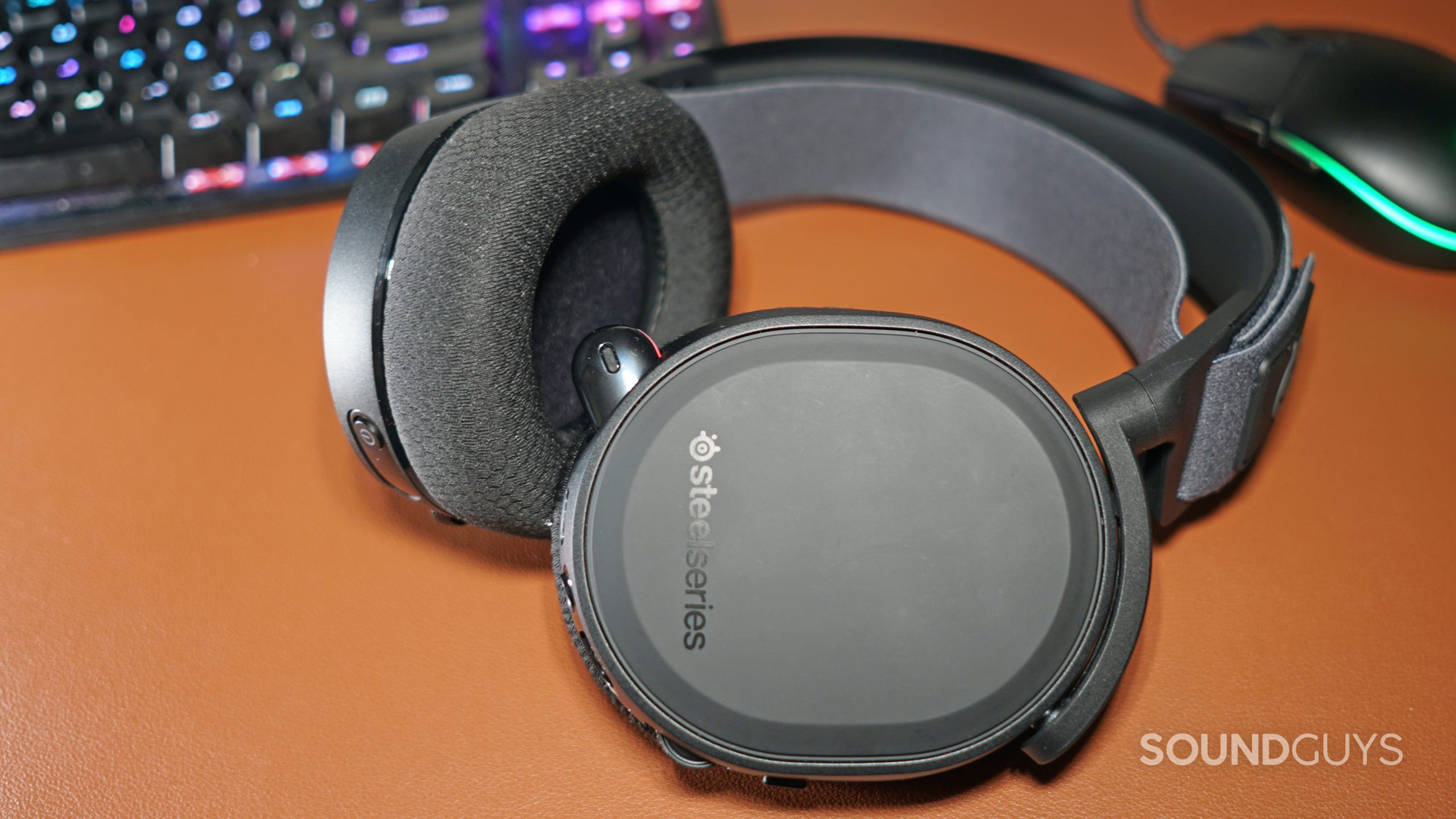The SteelSeries Arctis 7+ lays on a leather desk mat in front of a HyperX mechanical gaming keyboard and a Logitech gaming mouse.