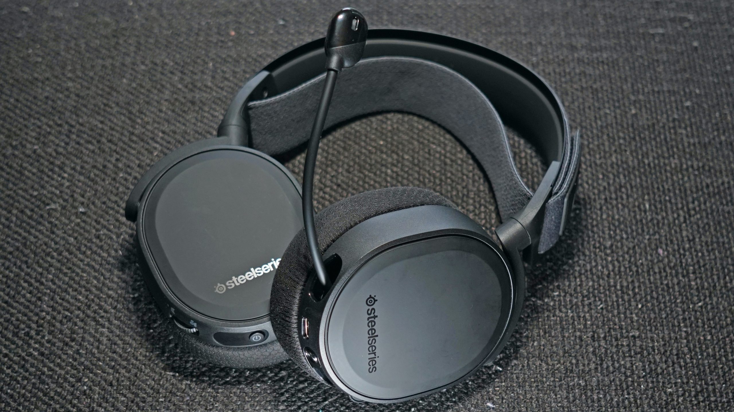 The SteelSeries Arctis 7+ lays on a fabric surface with its retractable microphone extended and