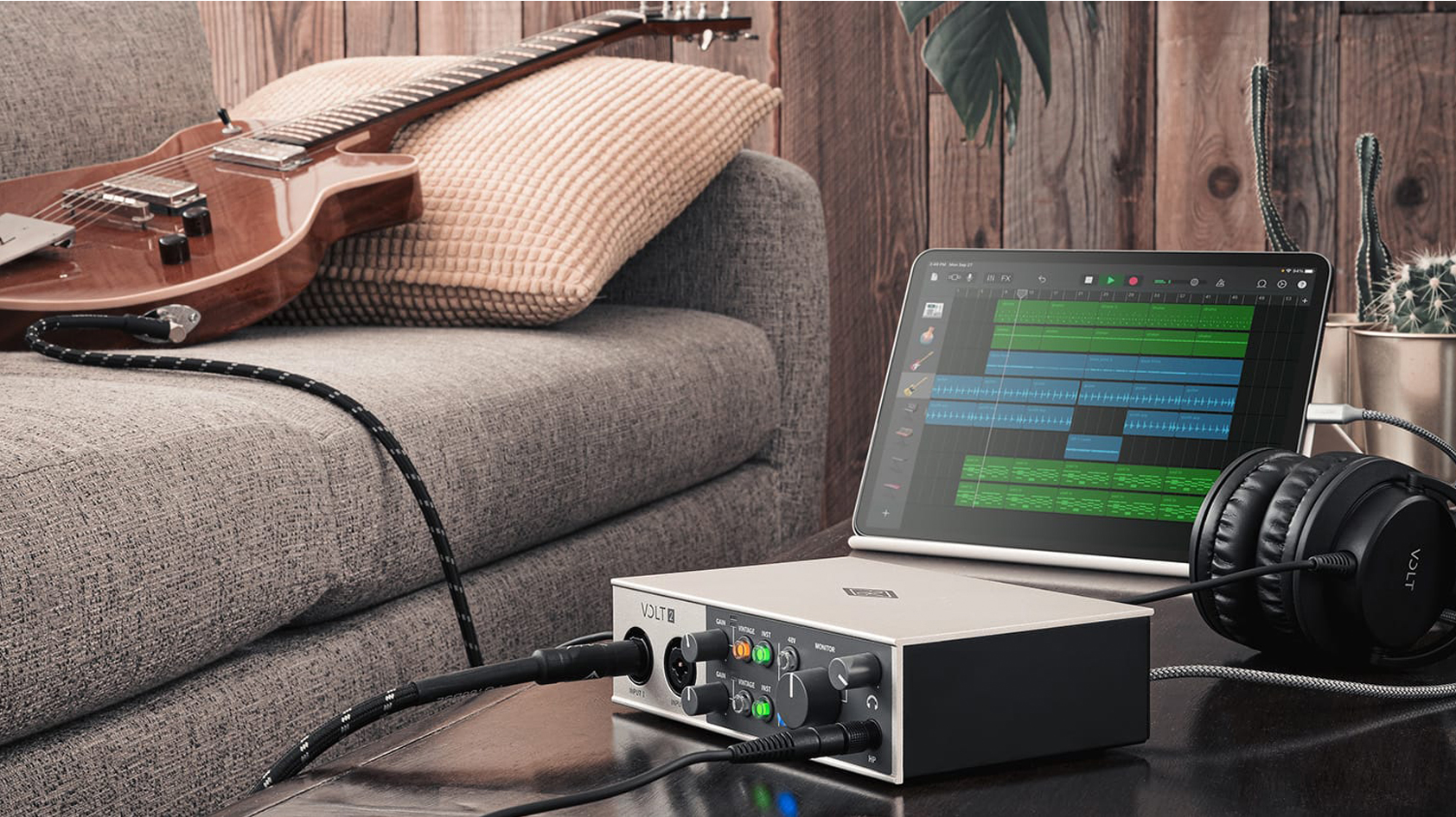 Promotional image of Universal Audio Volt 2 shown on a table plugged into an iPad with a guitar on a couch.