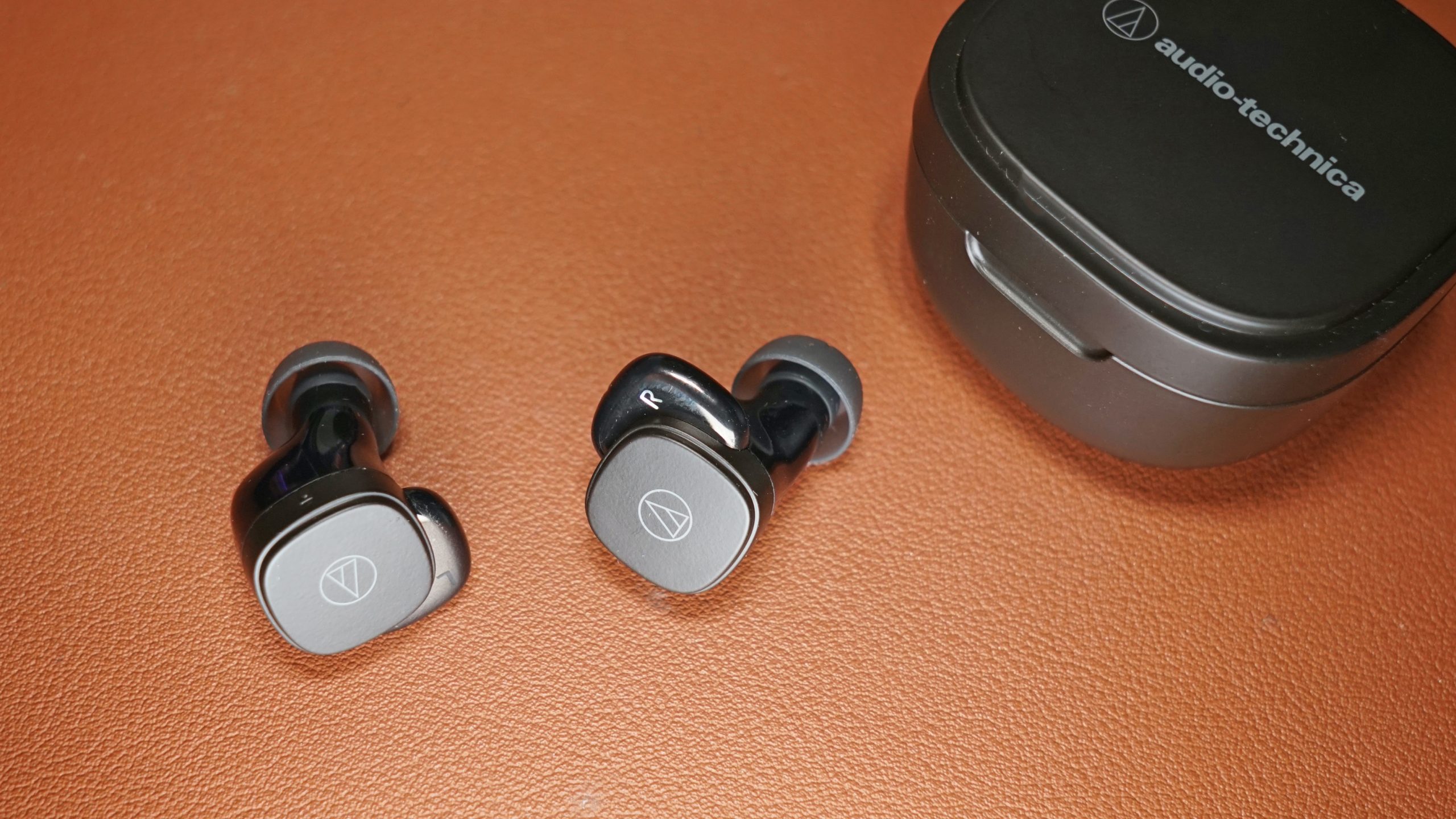 The Audio-Technica ATH-SQ1TW true wireless earbuds lay on a leather surface next to its charging case.