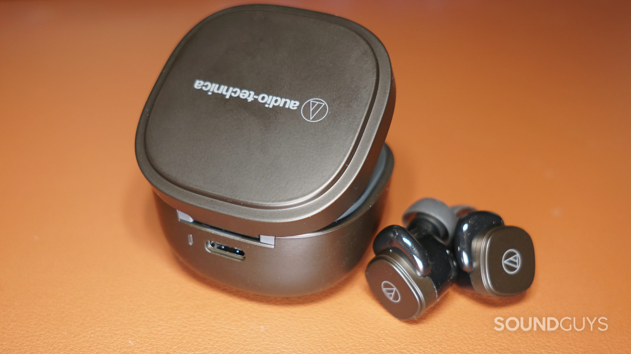The Audio-Technica ATH-SQ1TW charging case sits next to the earbuds on a leather surface with its USB-C charging port in view.