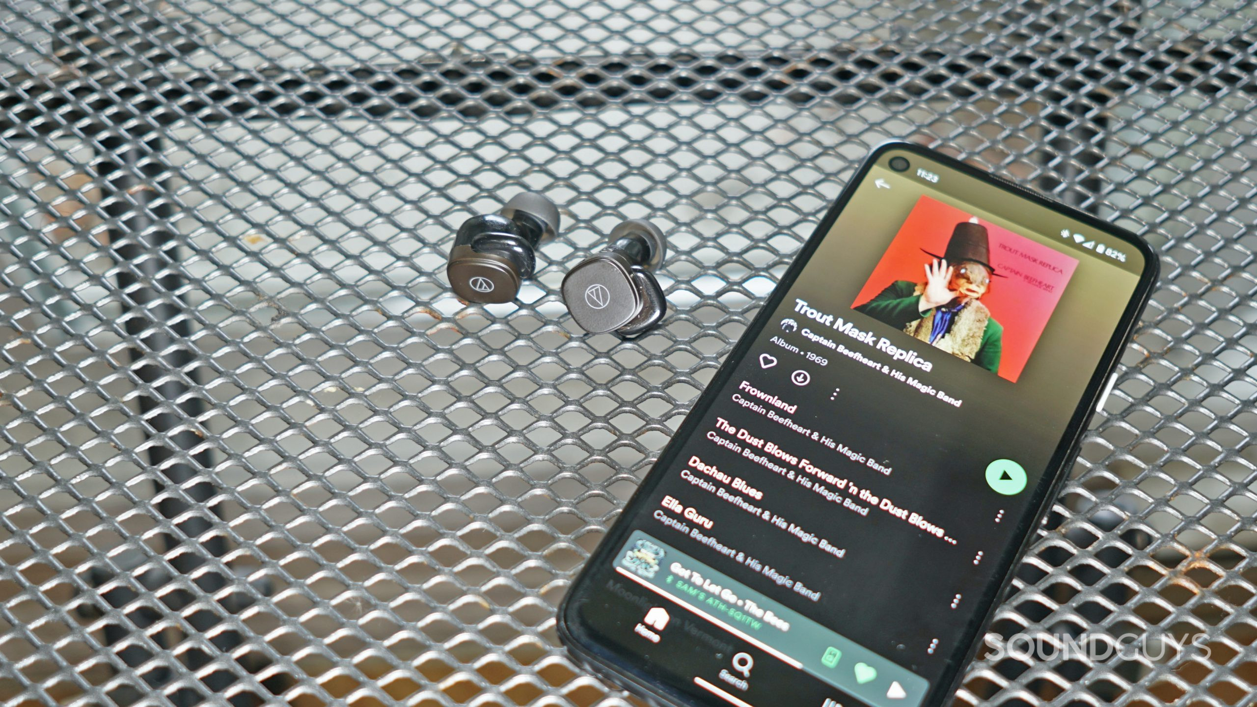 The Audio-Technica ATH-SQ1TW true wireless earbuds lay on a metal table next to a Google Pixel 4a playing Captain Beefheart