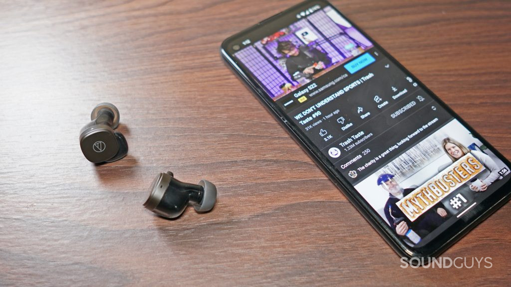 The Audio-Technica ATH-SQ1TW lays on a wooden table next to a Google Pixel 4a playing the Trash Taste podcast on YouTube