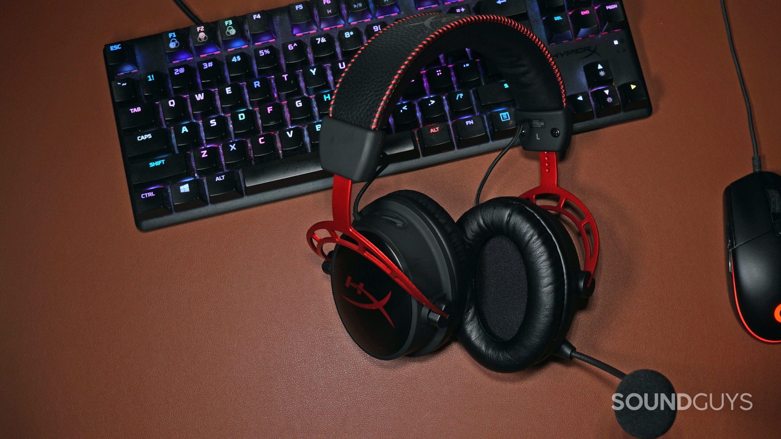 The HyperX Cloud Alpha Wireless gaming headset lays on a leather surface next to a HyperX mechanical gaming keyboard and a Logitech gaming mouse.