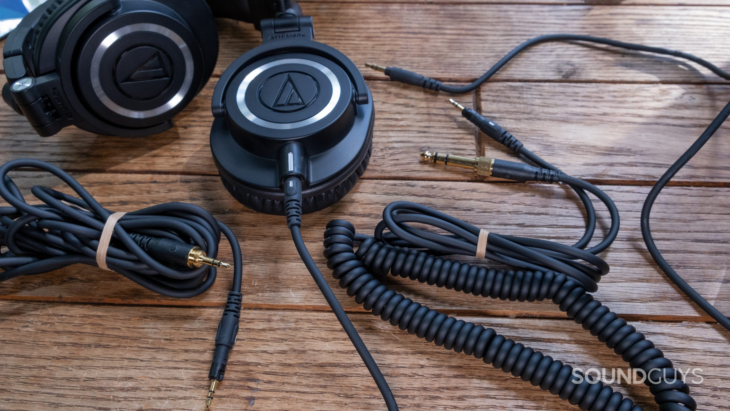 The Audio-Technica ATH-M50x lays partially folded on a wood surface surrounded by all three included cables.