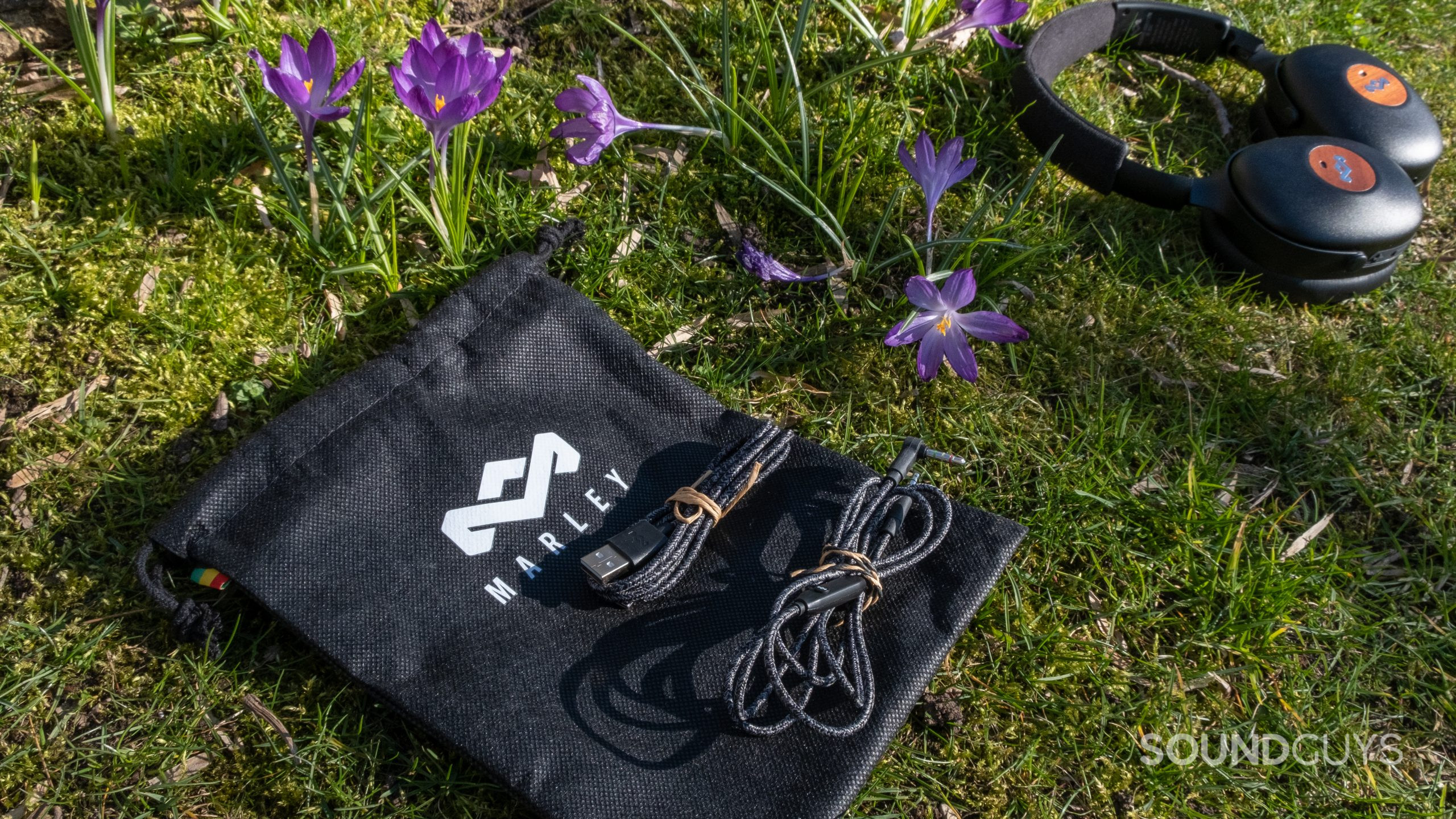 The accessories of the House of Marley Positive Vibration XL ANC including a carry pouch, headphone cable, and USB-C charging cable lay on grass next to flowers.