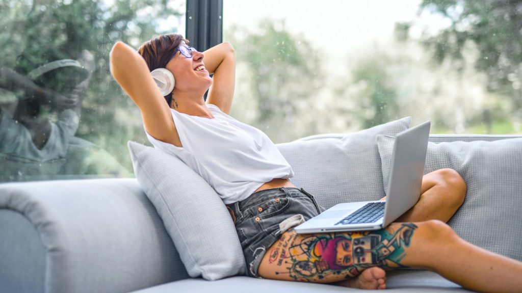 A woman relaxes on a couch while listening to music on wireless headphones through a laptop.