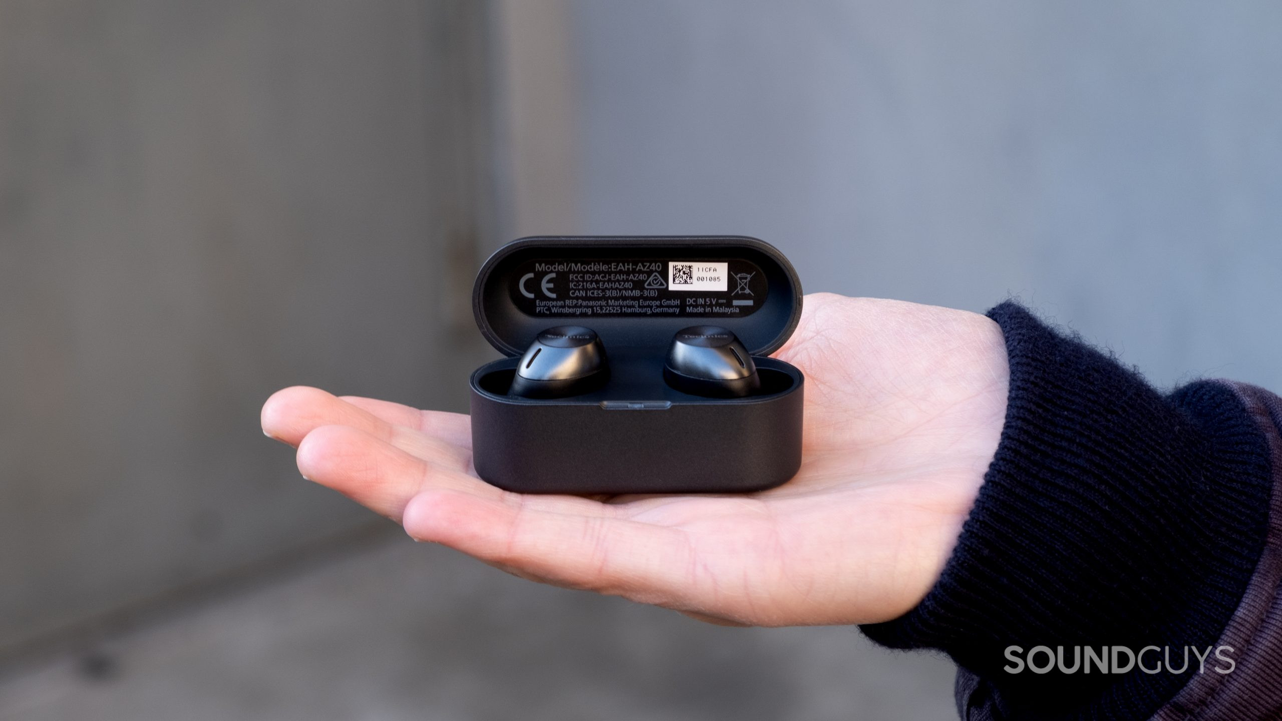 A hand holds the Technics EAH-AZ40 in the palm with the case open with a concrete background.