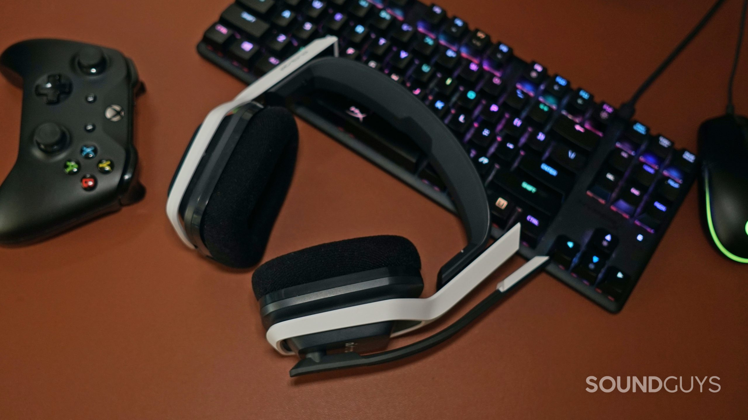 The Astro A20 gaming headset lays on a leather surface in front of a HyperX gaming keyboard, a Logitech gaming mouse, and a Microsoft Xbox controller.