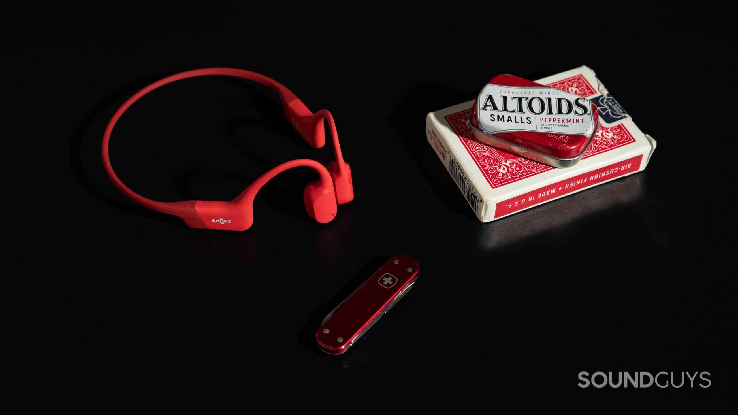 The Shokz OpenRun bone conduction headphones in red next to a pack of cards, mini Altoids tin, and mini Swiss Army Knife.