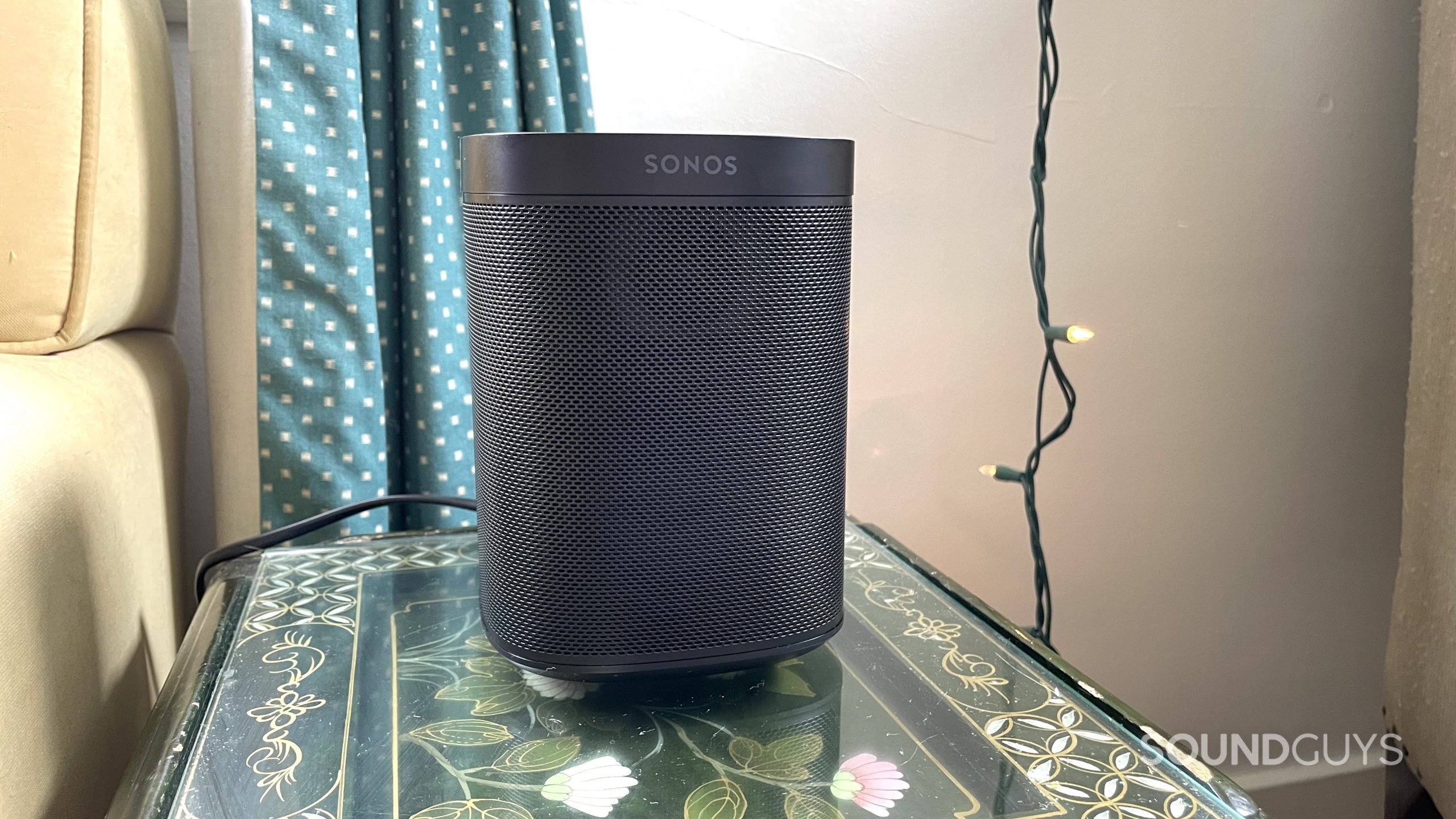 Sonos One Gen 2) on a side table with curtains and string lights in the background.