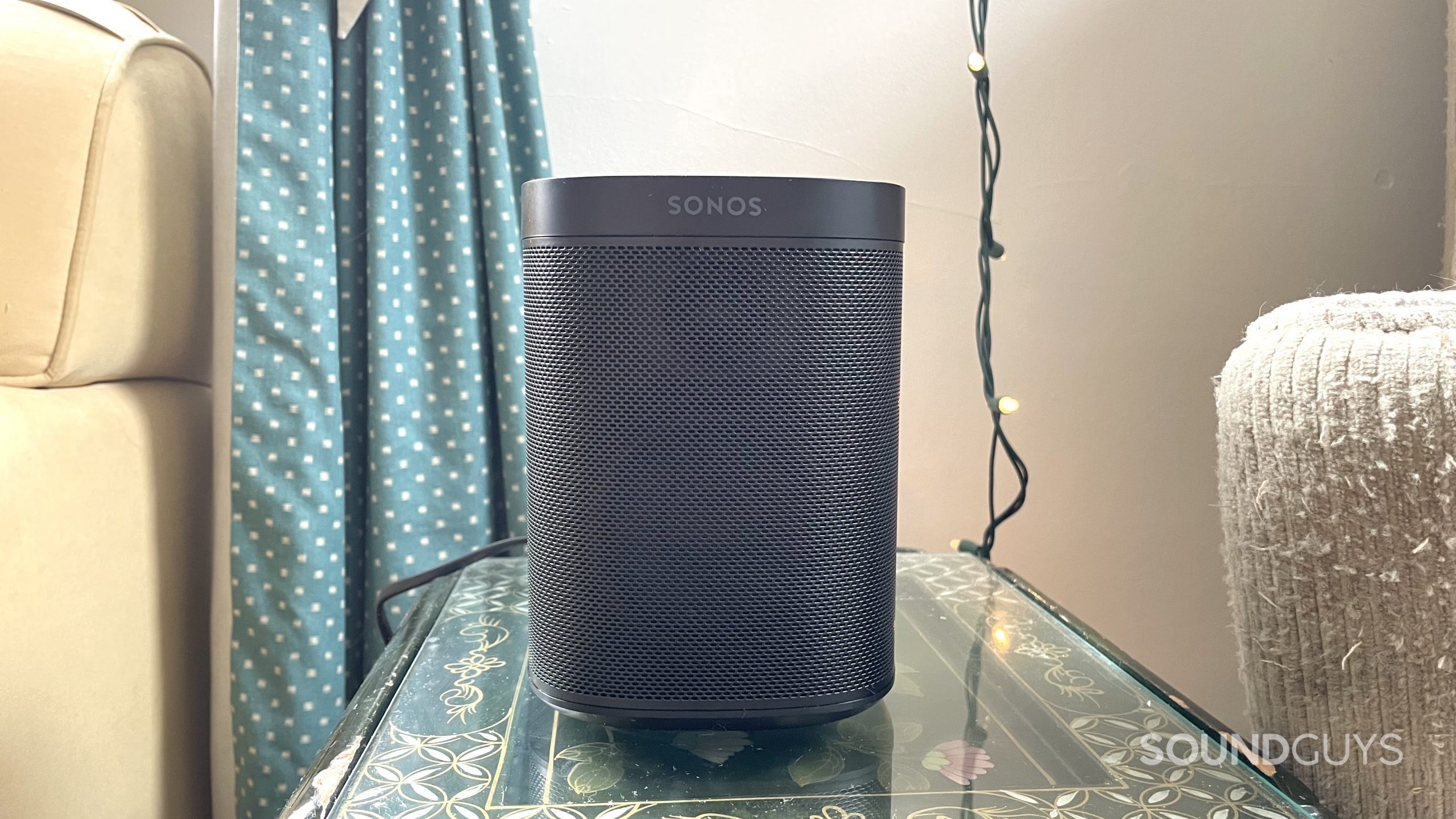 Sonos One Gen 2) on a side table with curtains and string lights in the background.