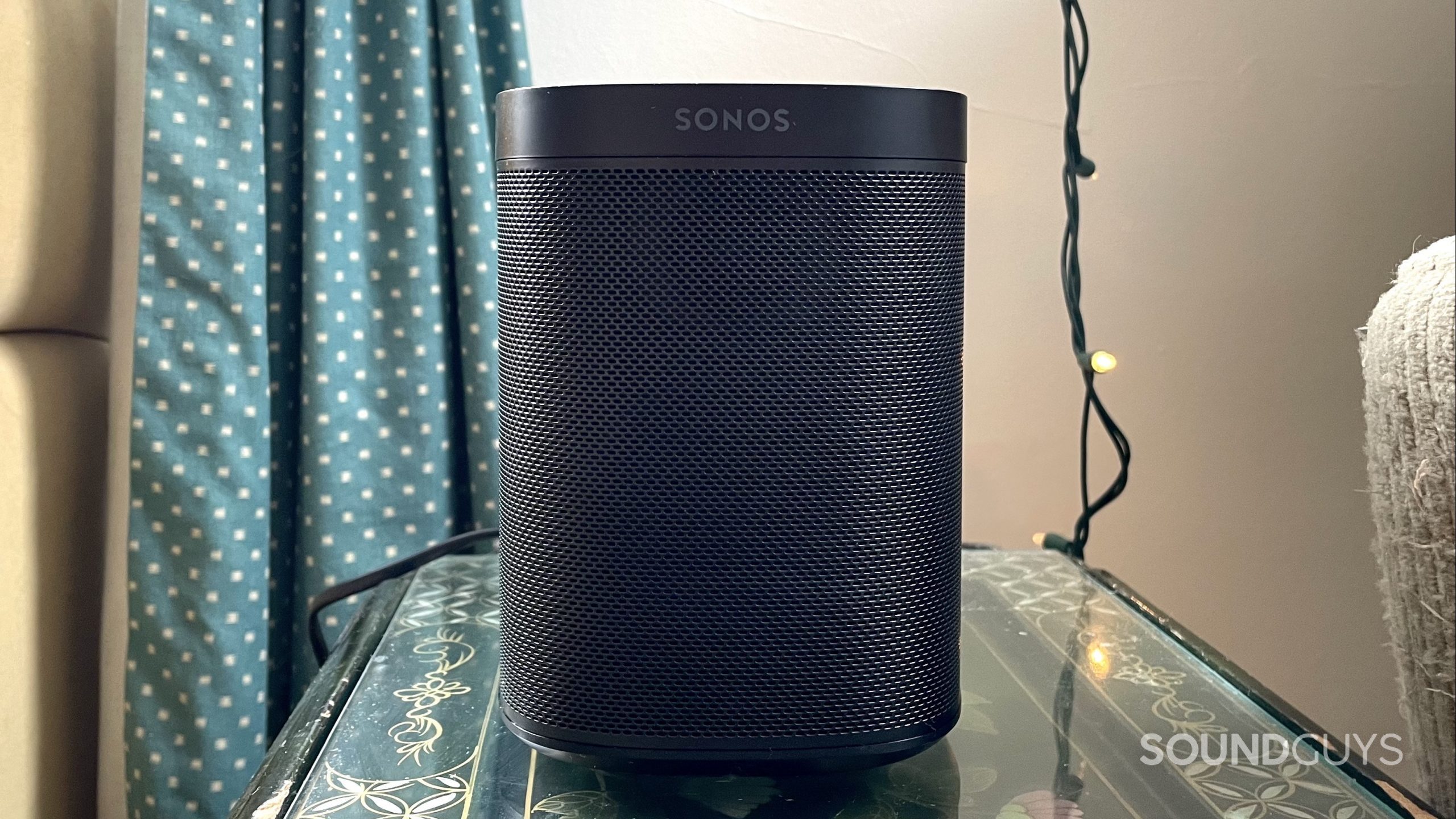 Sonos One (Gen 2) on a side table with curtains and string lights in the background.