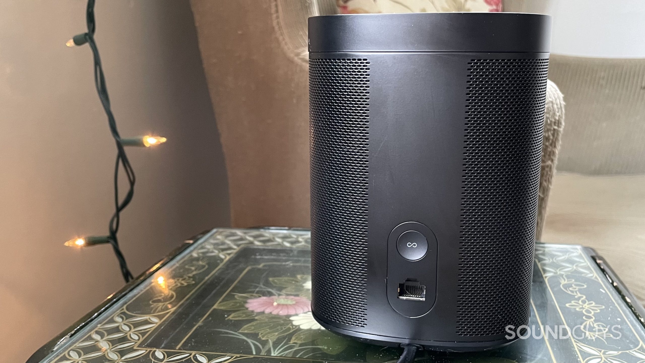 Back of the Sonos One Gen 2) on a side table with string lights and an armchair in the background.