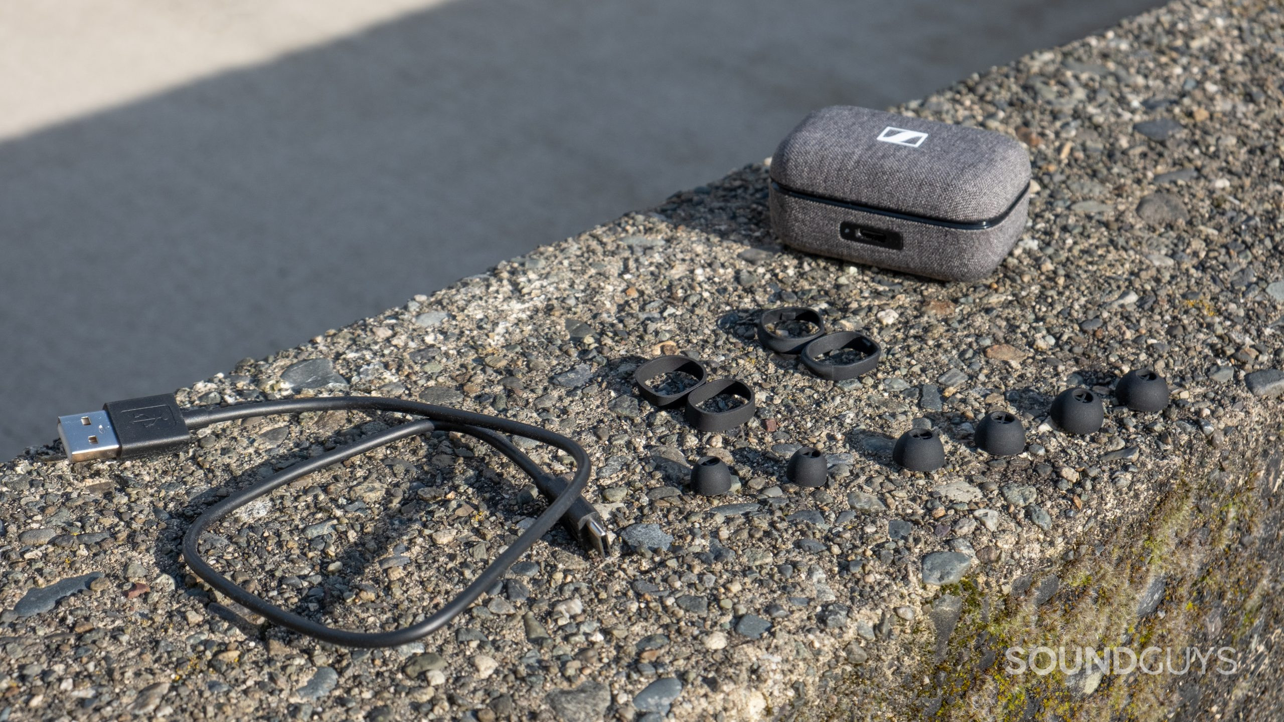 The ear tips, case, and stabilizers included with the Sennheiser MOMENTUM True Wireless 3 displayed on mossy concrete.