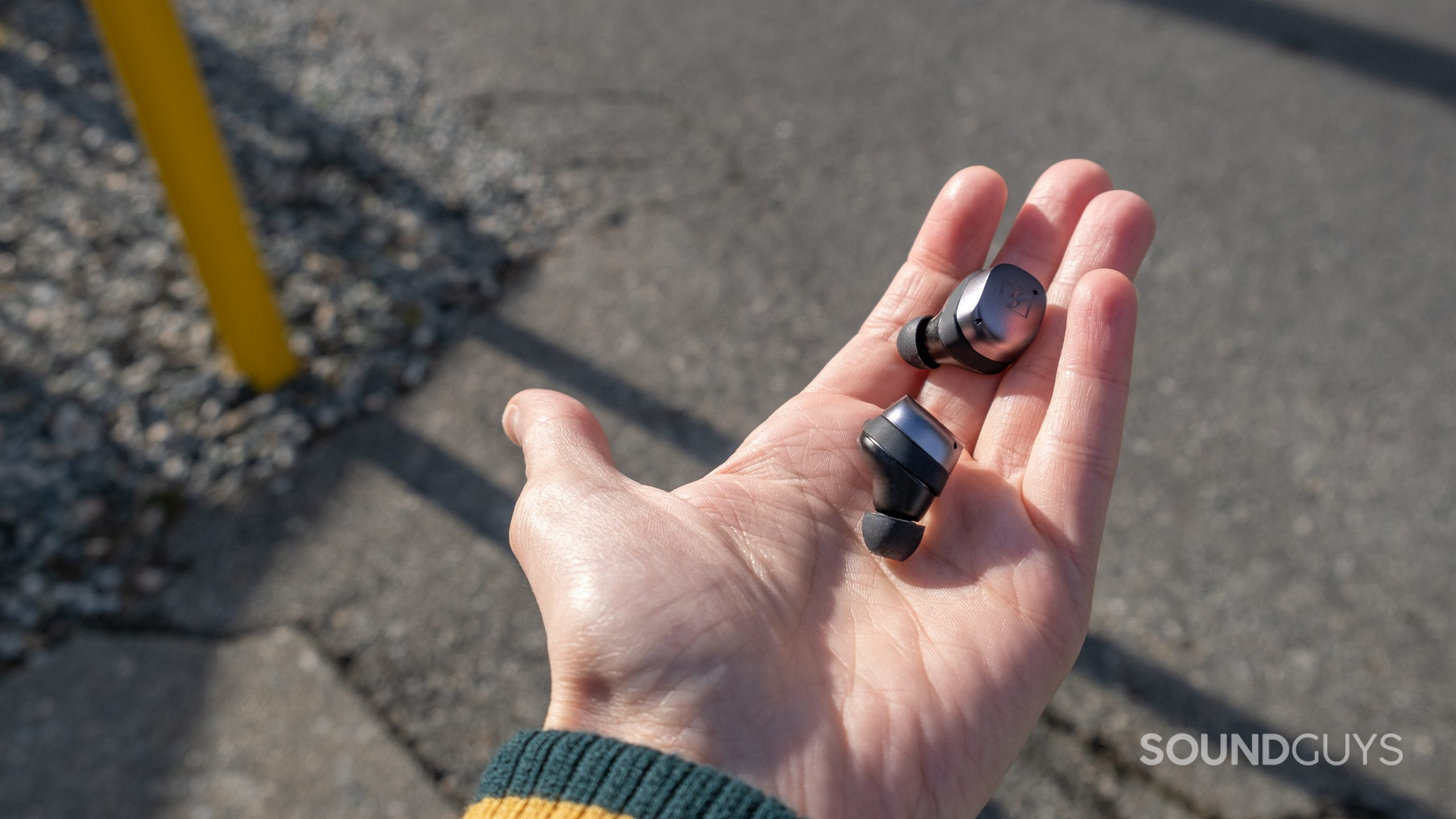 A hand holds the Sennheiser MOMENTUM True Wireless 3 earbuds above concrete.