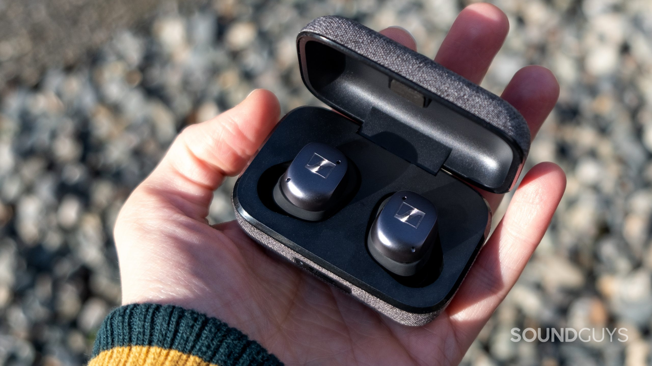 A hand holds the open case of the Sennheiser MOMENTUM True Wireless 3 with blurred rocks in the background.