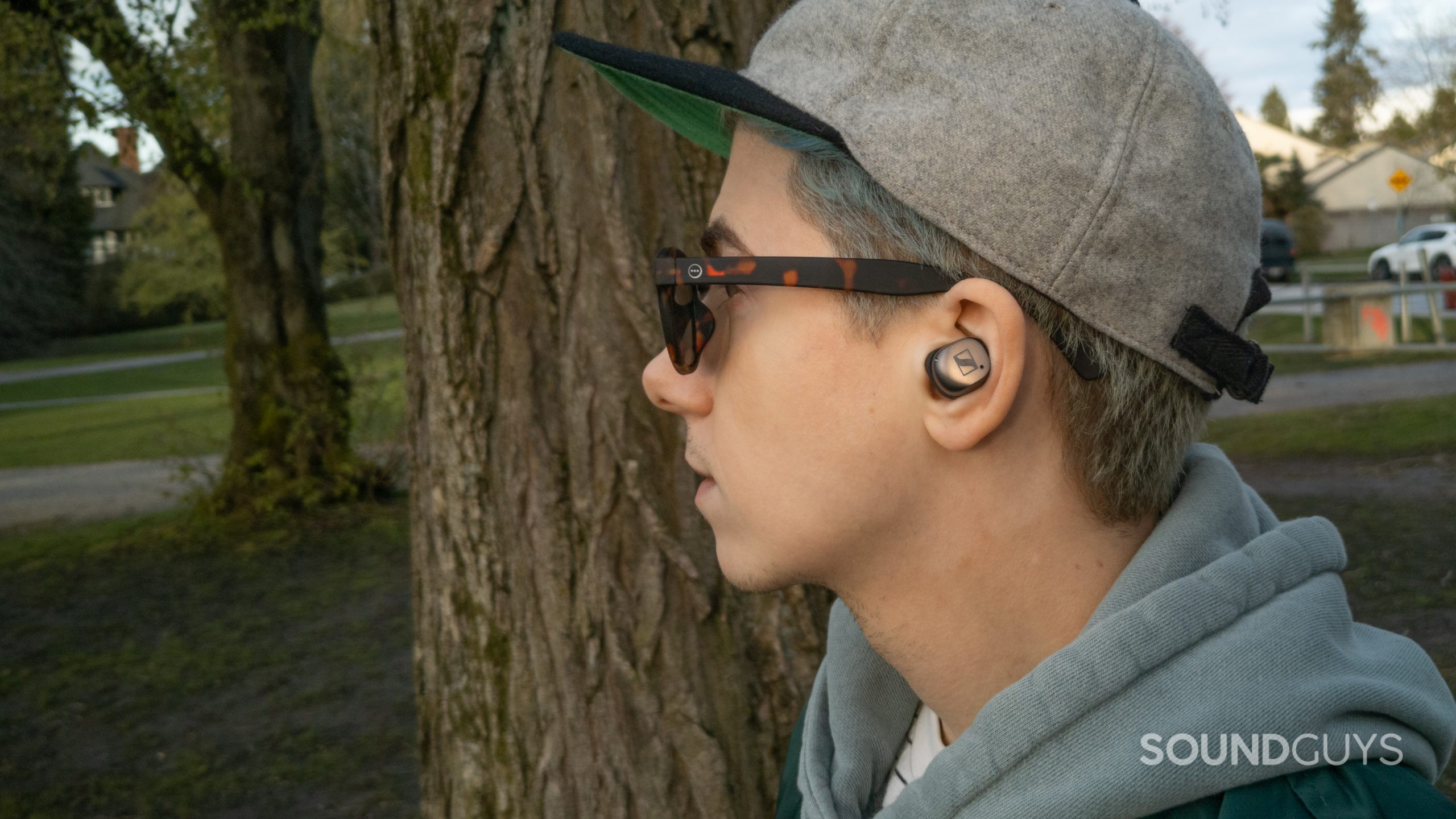 A man faces left wearing the Sennheiser MOMENTUM True Wireless 3 in front of trees and grass near a parking lot.
