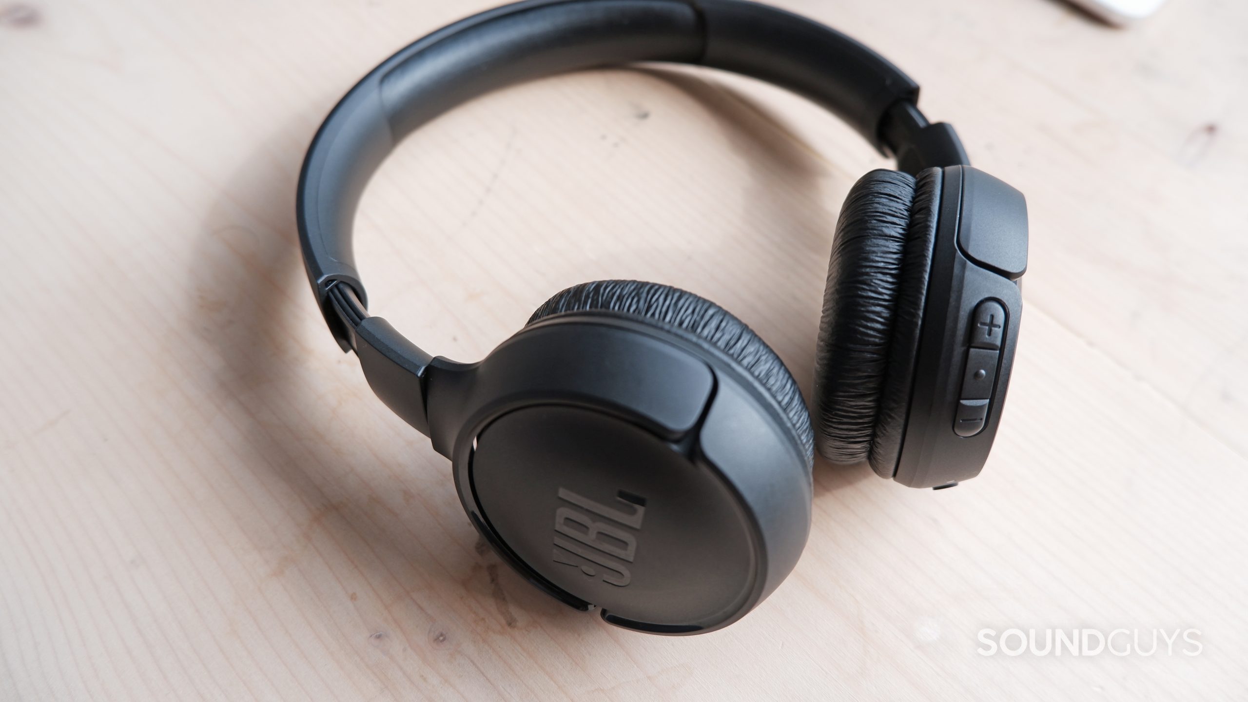 The JBL Tune 510BT headset on a wooden table, showing the volume buttons and ear cups.