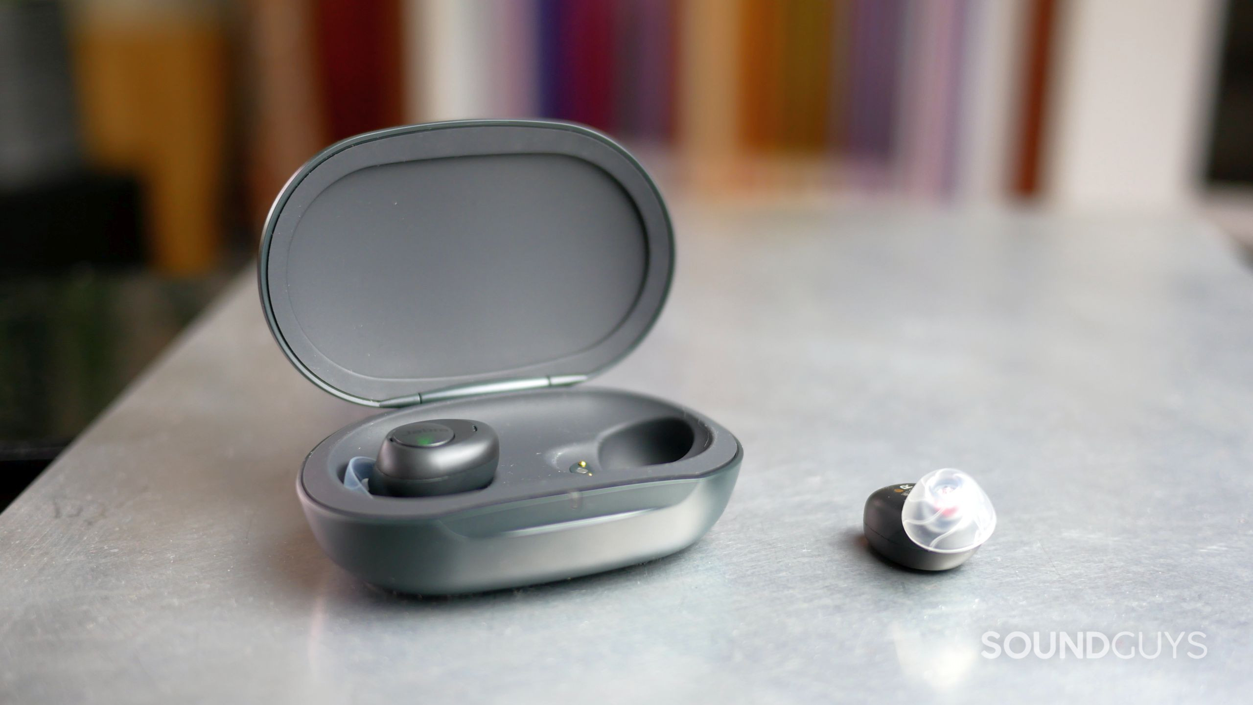 Jabra Enhance Plus open charging case with one earbud and a second lying next to it.