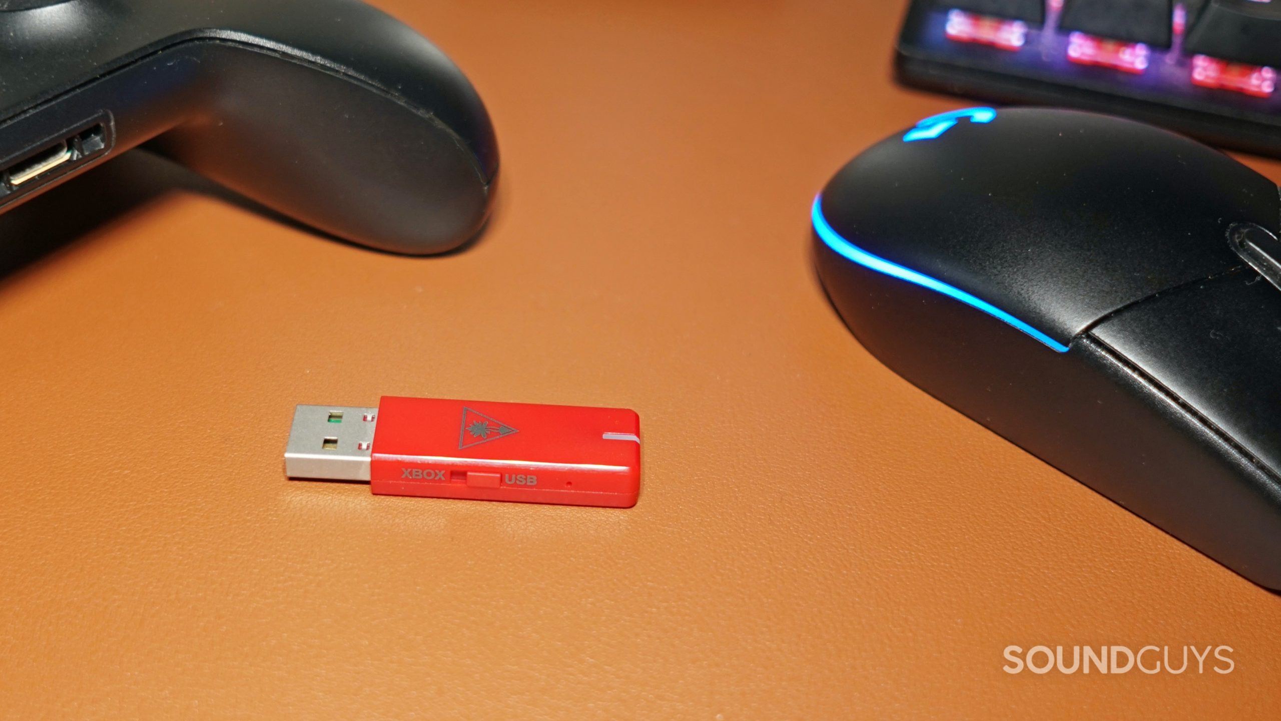 The Turtle Beach Stealth 600 Gen 2 MAX usb dongle lays on a leather surface next to an Xbox One controller and a Logitech Gaming Mouse