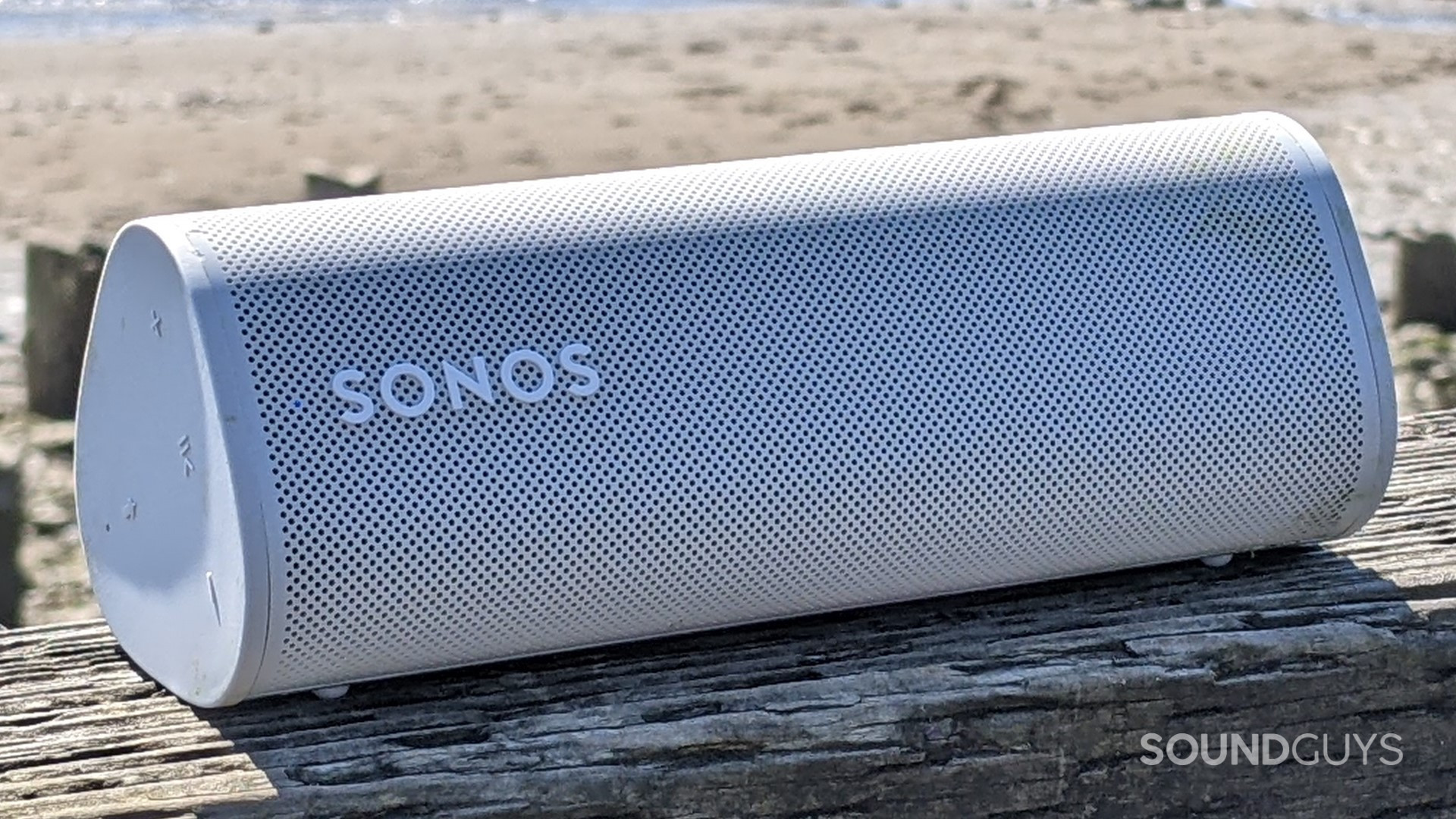 A white Sonos Roam sitting on a wood raining in front of a beach on a sunny day.