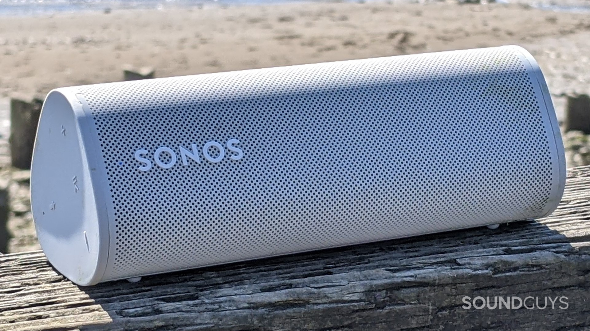 A white Sonos Roam sitting on a wooden railing in front of a beach on a sunny day.