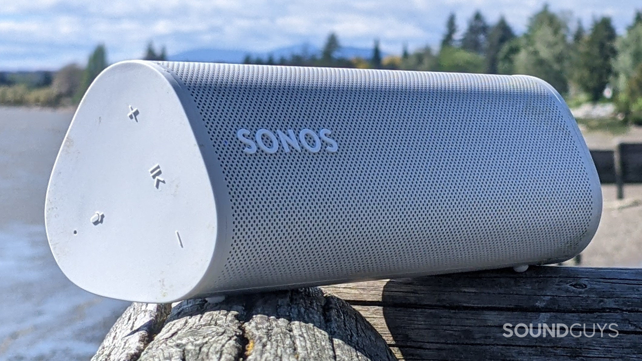 A white Sonos Roam speaker sitting diagonally across the corner of a wooden railing in front of a body of water and some trees surrounding a beach in the distant background.