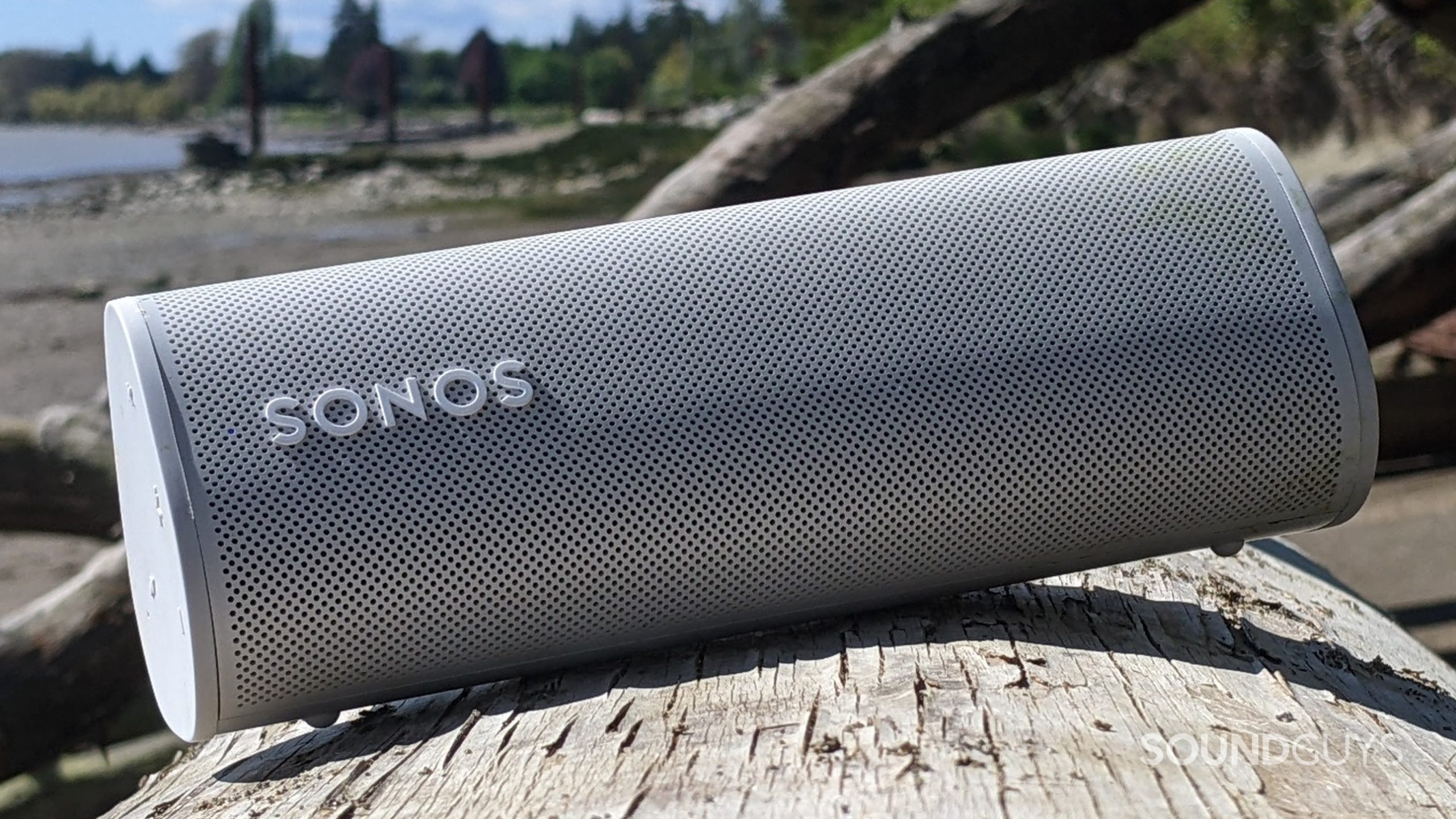 A white Sonos Roam speaker sitting at a slight angle on a log on a beach in front of a fallen tree and a body of water along with some trees in the distance.