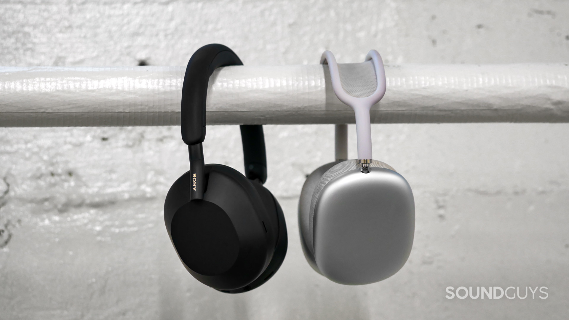 Side-on view of Sony WH-1000XM5 beside the AirPods Max hanging over a white horizontal pipe