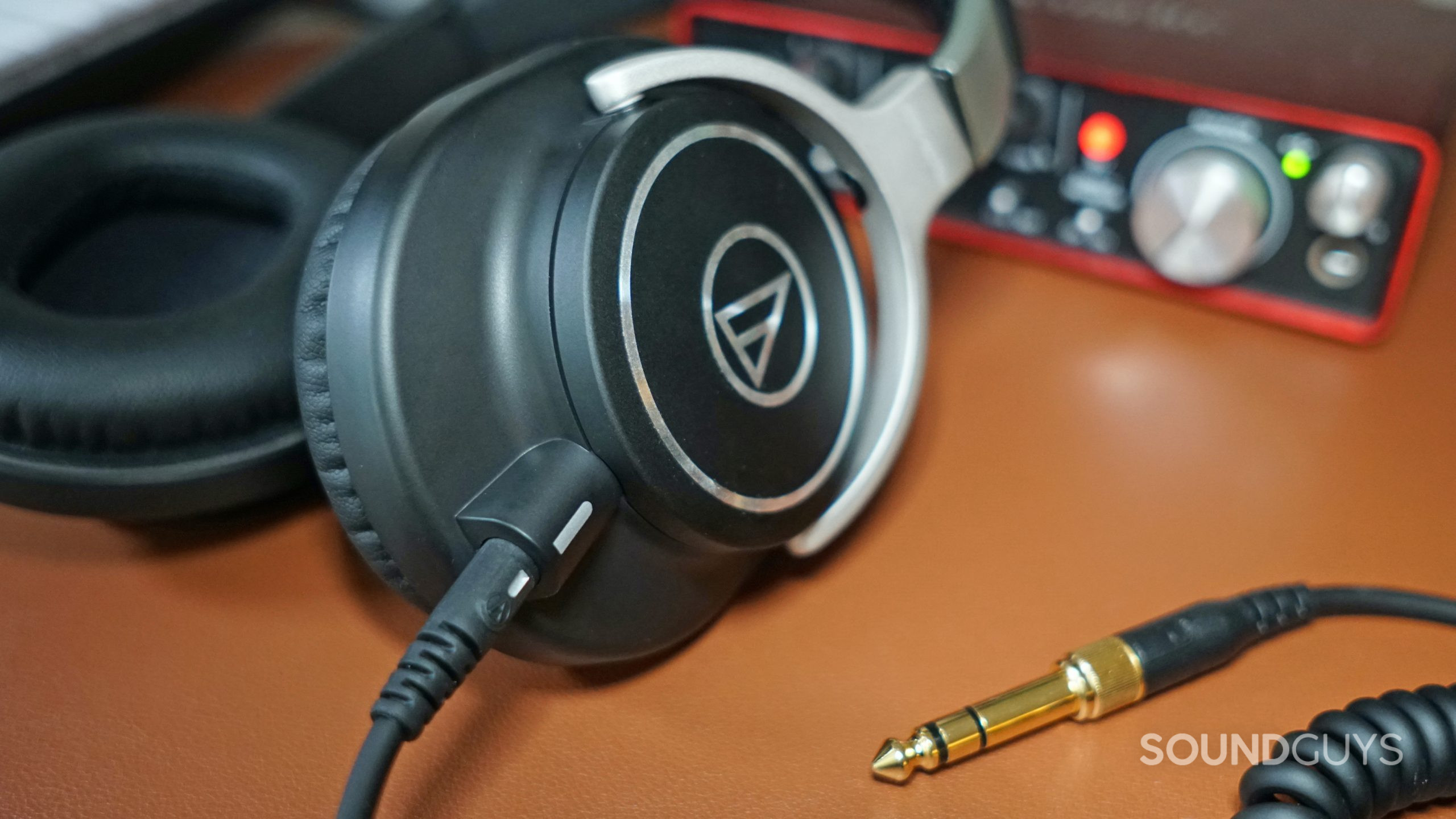 The Audio-Technica ATH-M70x lays on a leather surface in front of a Focusrite Scarlett 2i2, with its 1/4 inch plug on the table.