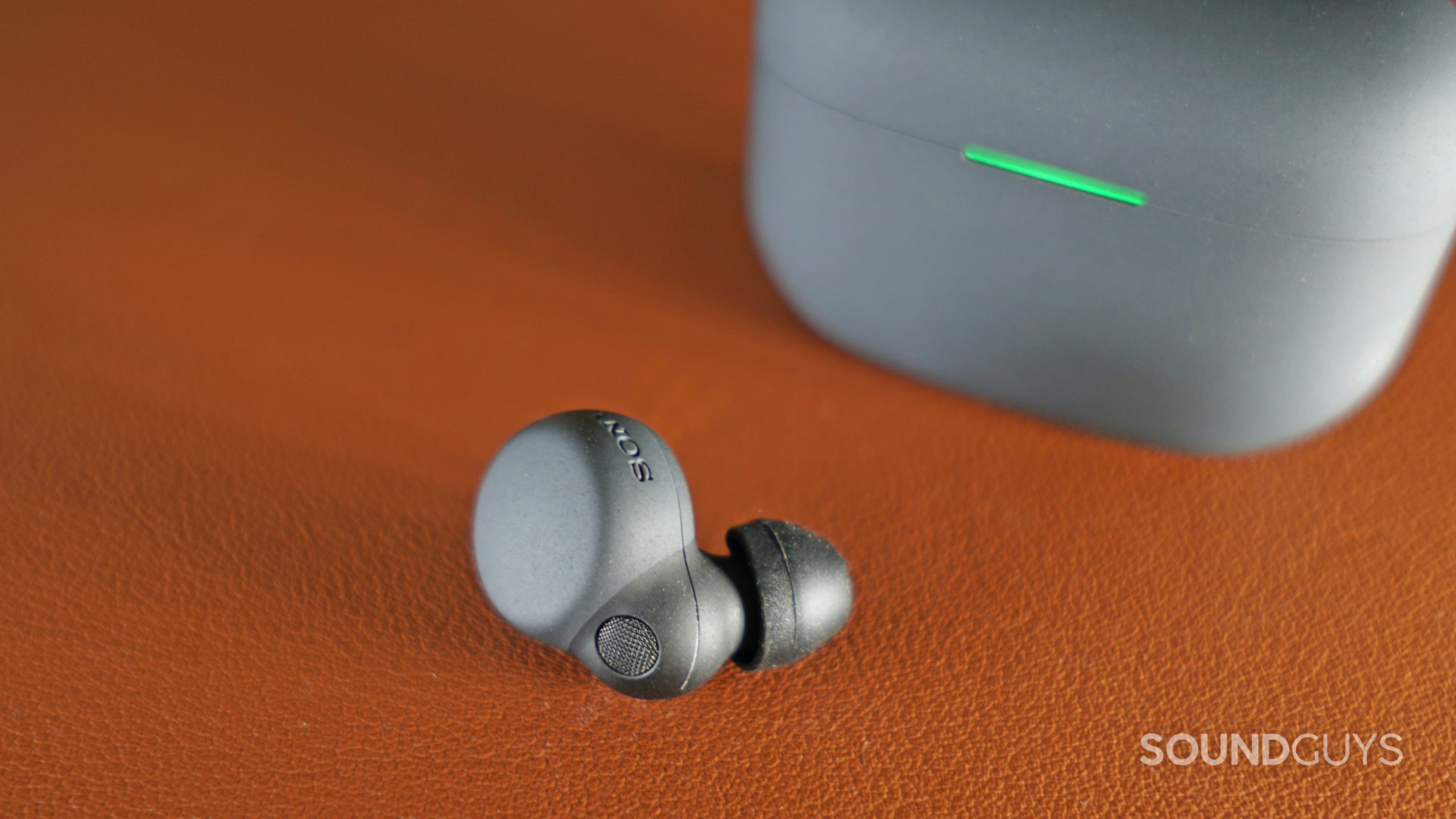 The Sonly LinkBuds S earbud lays on a leather surface in front of its charging case.