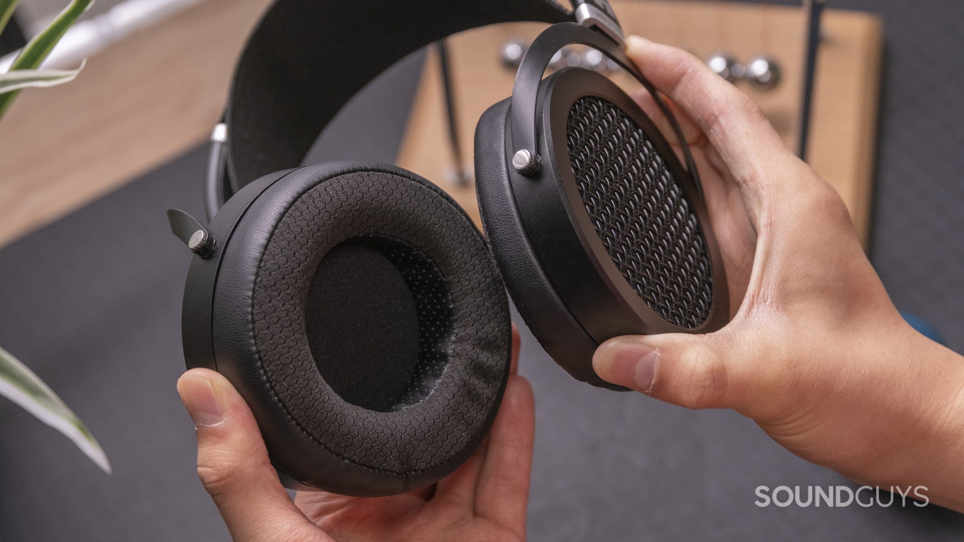 Hands hold the HiFiMan Sundara open-back headphones in front of a black backdrop with wooden accessories.