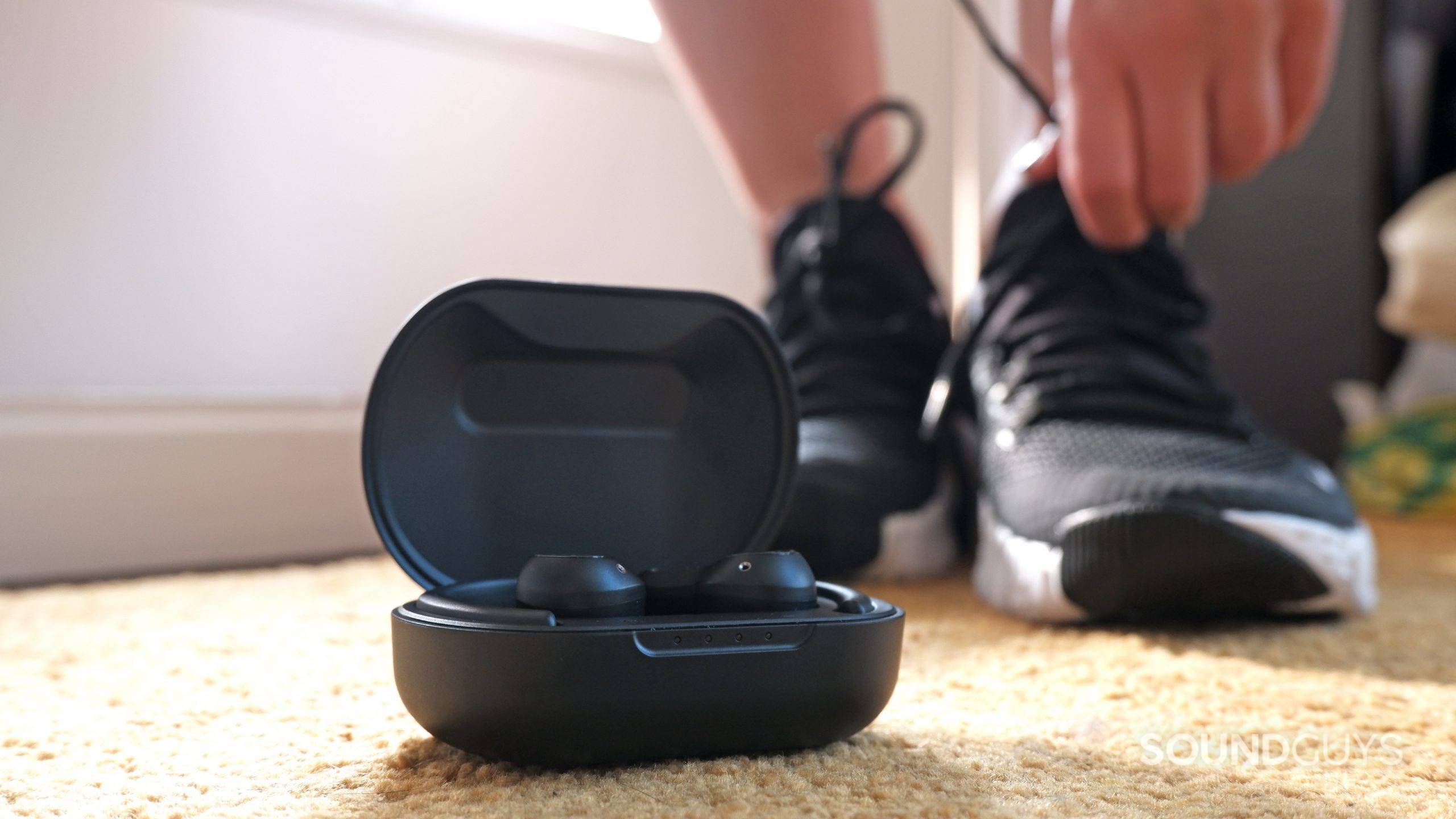 The JLab Epic Air Sport ANC in their case on the floor, with a person putting on running shoes in the background.