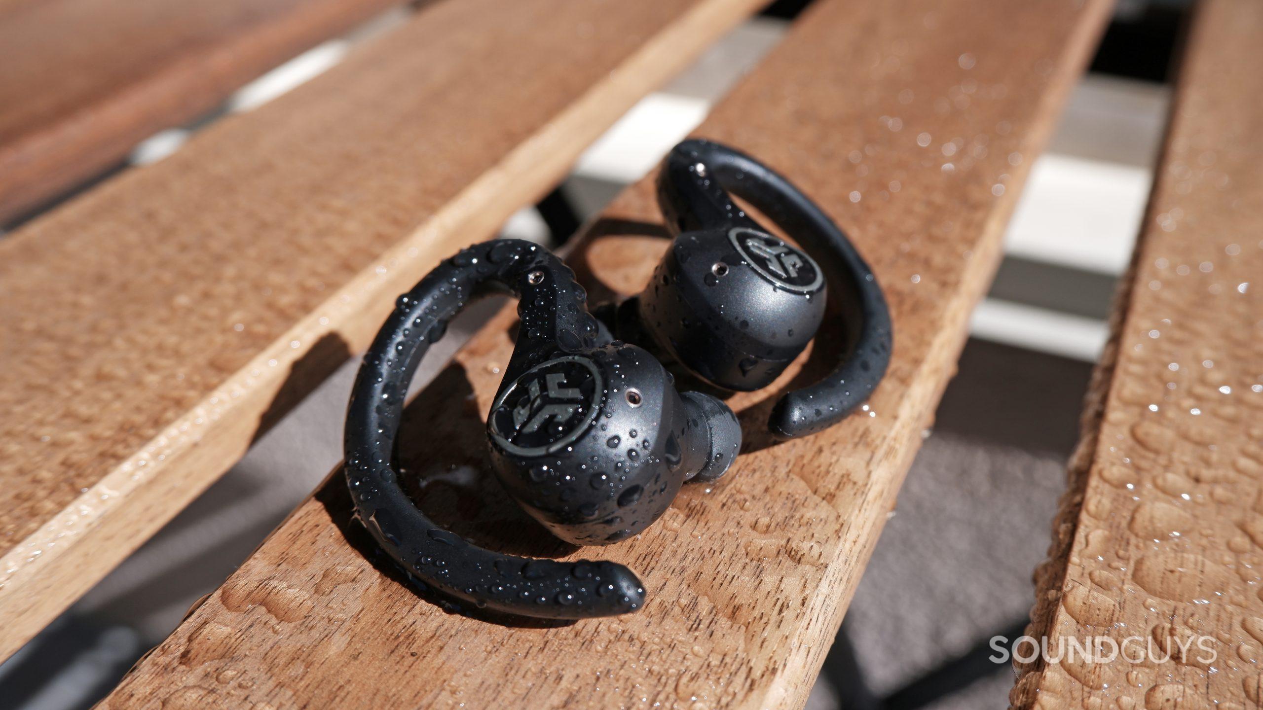 The JLab Epic Air Sport ANC on a wooden table ouside, with water droplets on the earbuds and table.