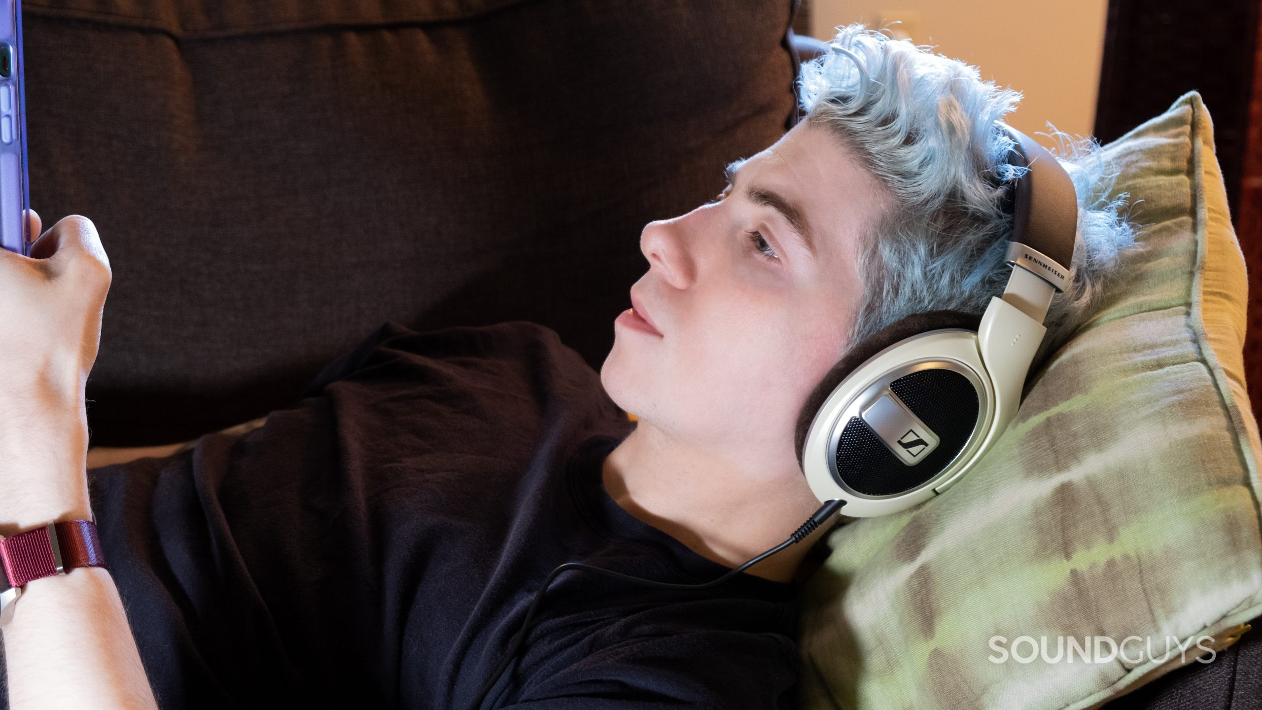 A man on a couch rests his head on a pillow while wearing the Sennheiser HD 599 and looking at a phone.