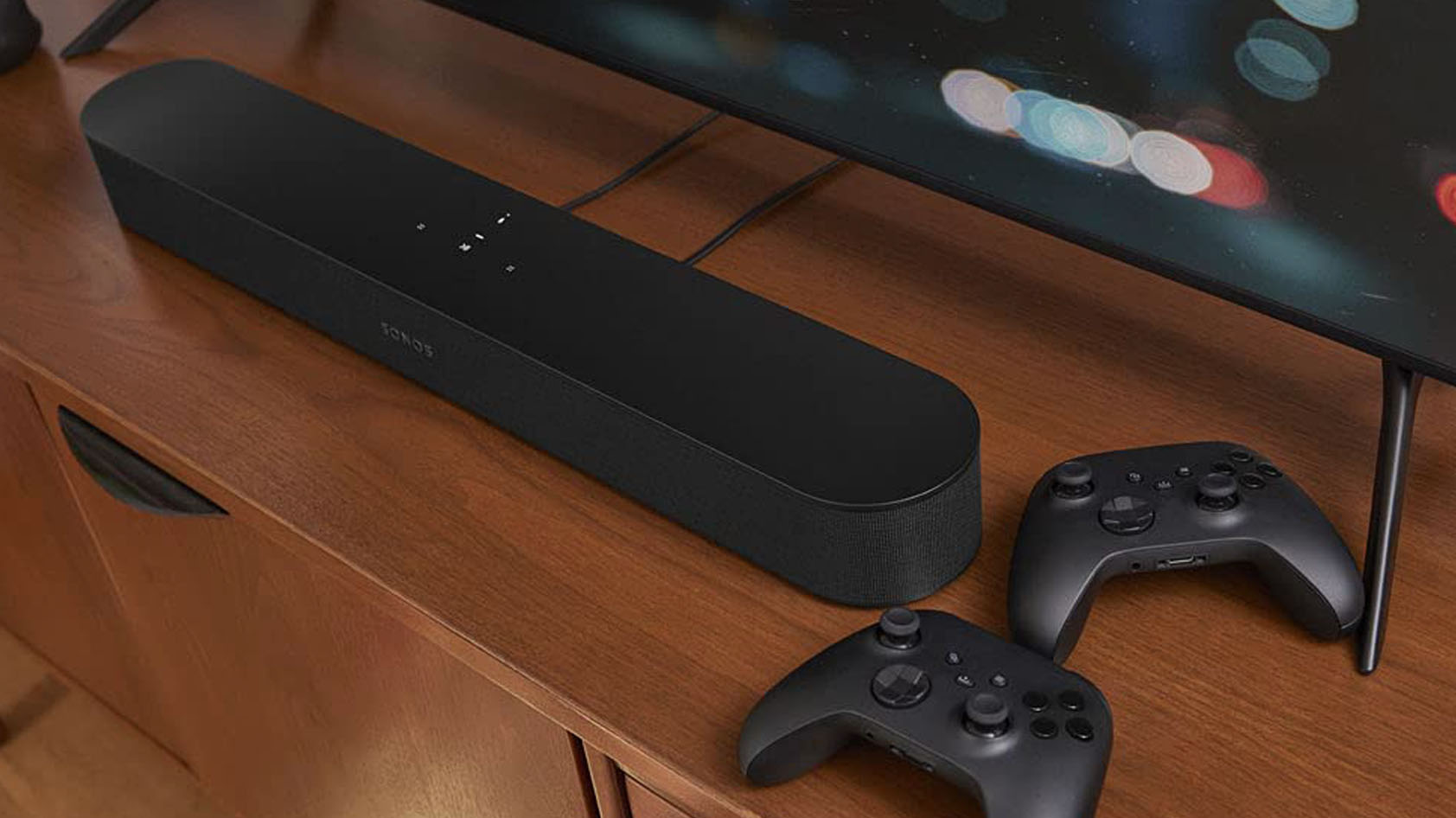The Sonos Beam (Gen 2) soundbar rests on a wooden TV stand next to two Xbox controllers.