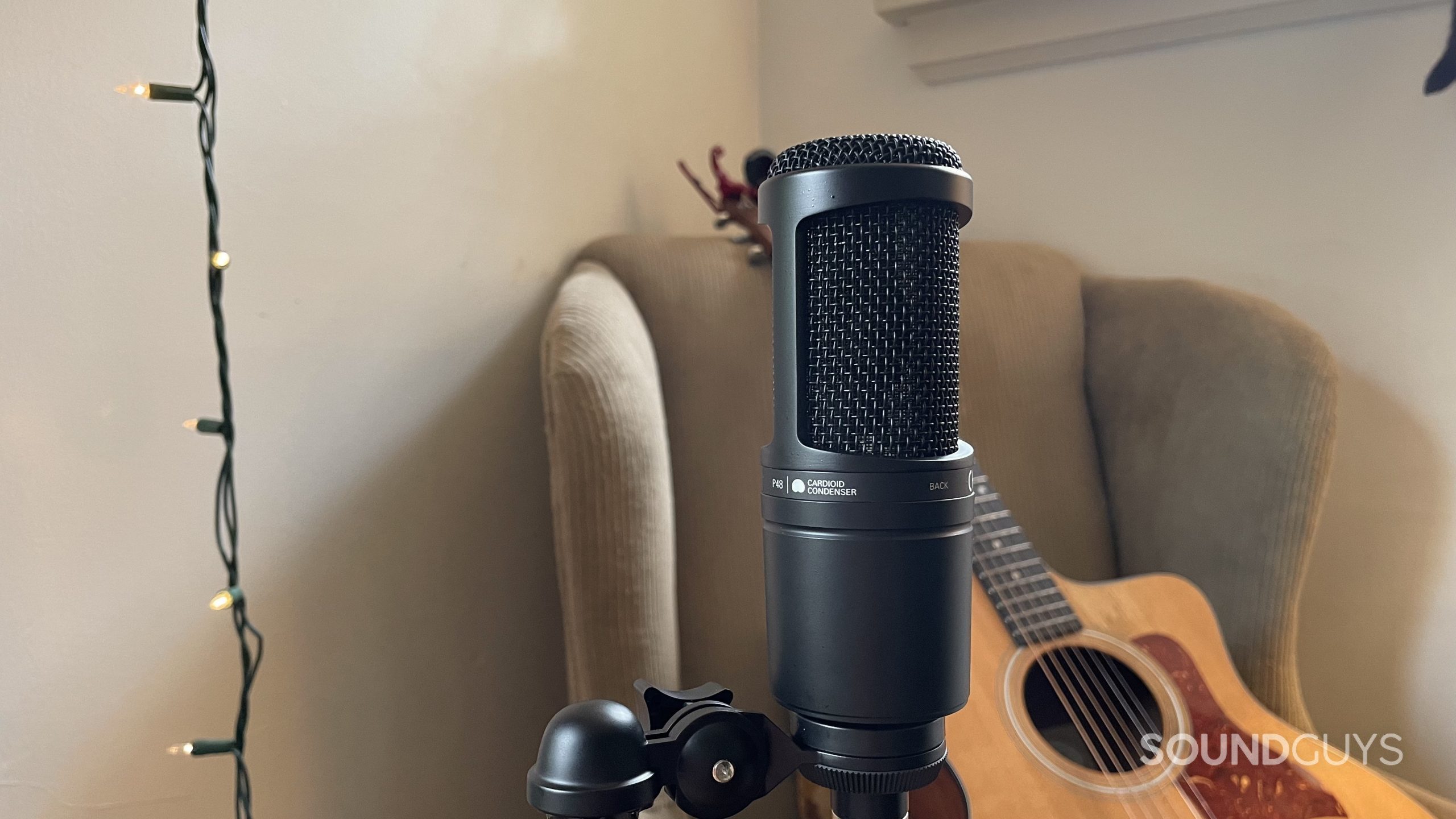 Audio-Technica AT2020 with a string of lights and an acoustic guitar resting on an armchair in the background.
